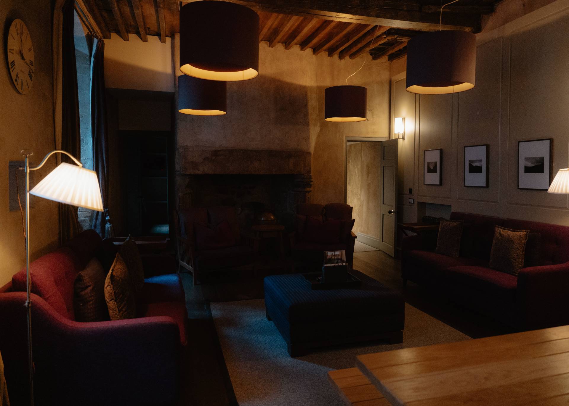 Red sofas and armchairs round a low table before a stone fireplace in a wood-beamed room, pendant lamps lit above, a clock and framed prints on the wall.