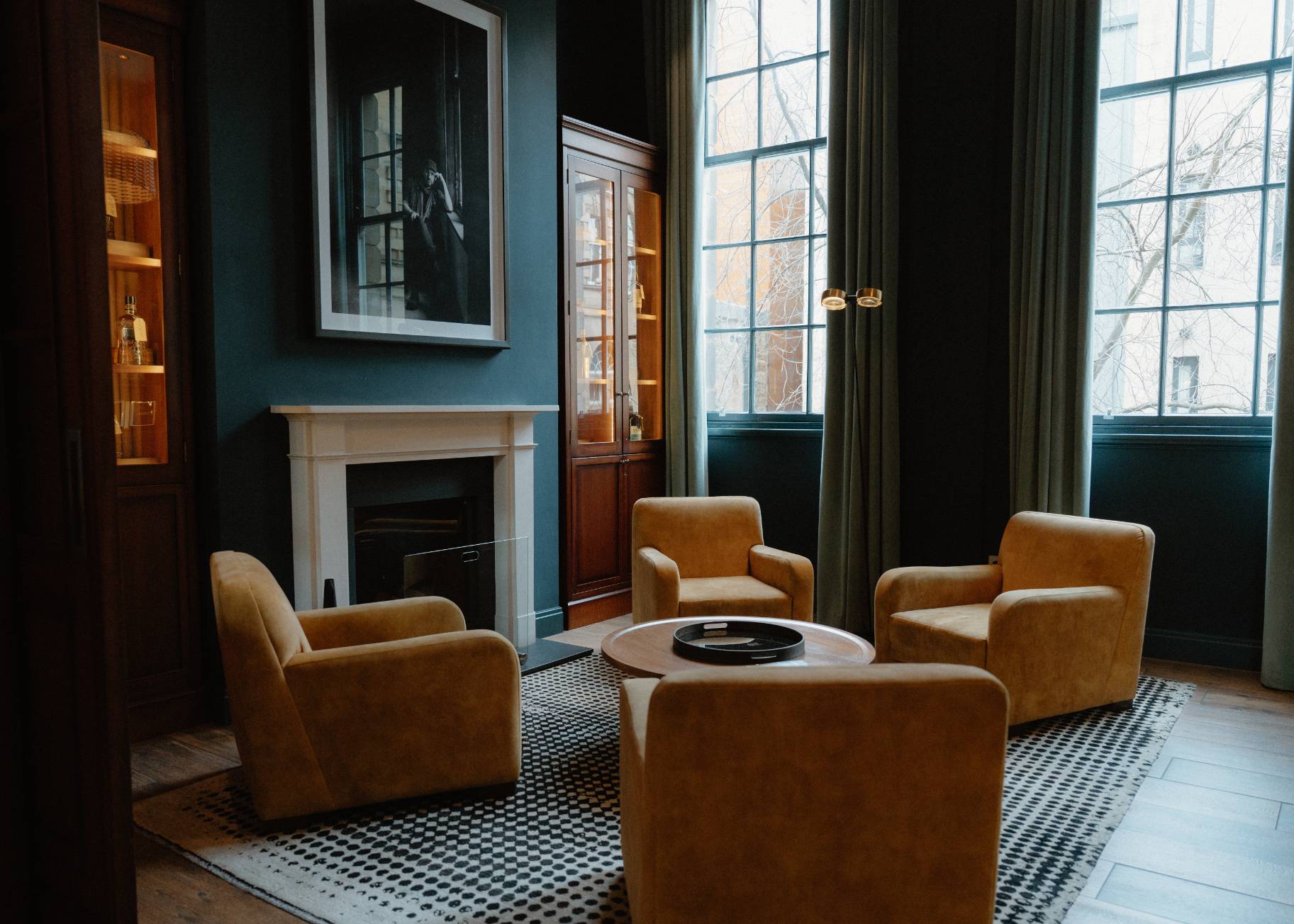 Mustard armchairs surround a round table before a fireplace with a framed black and white portrait, glass-fronted cabinets flanking the room.