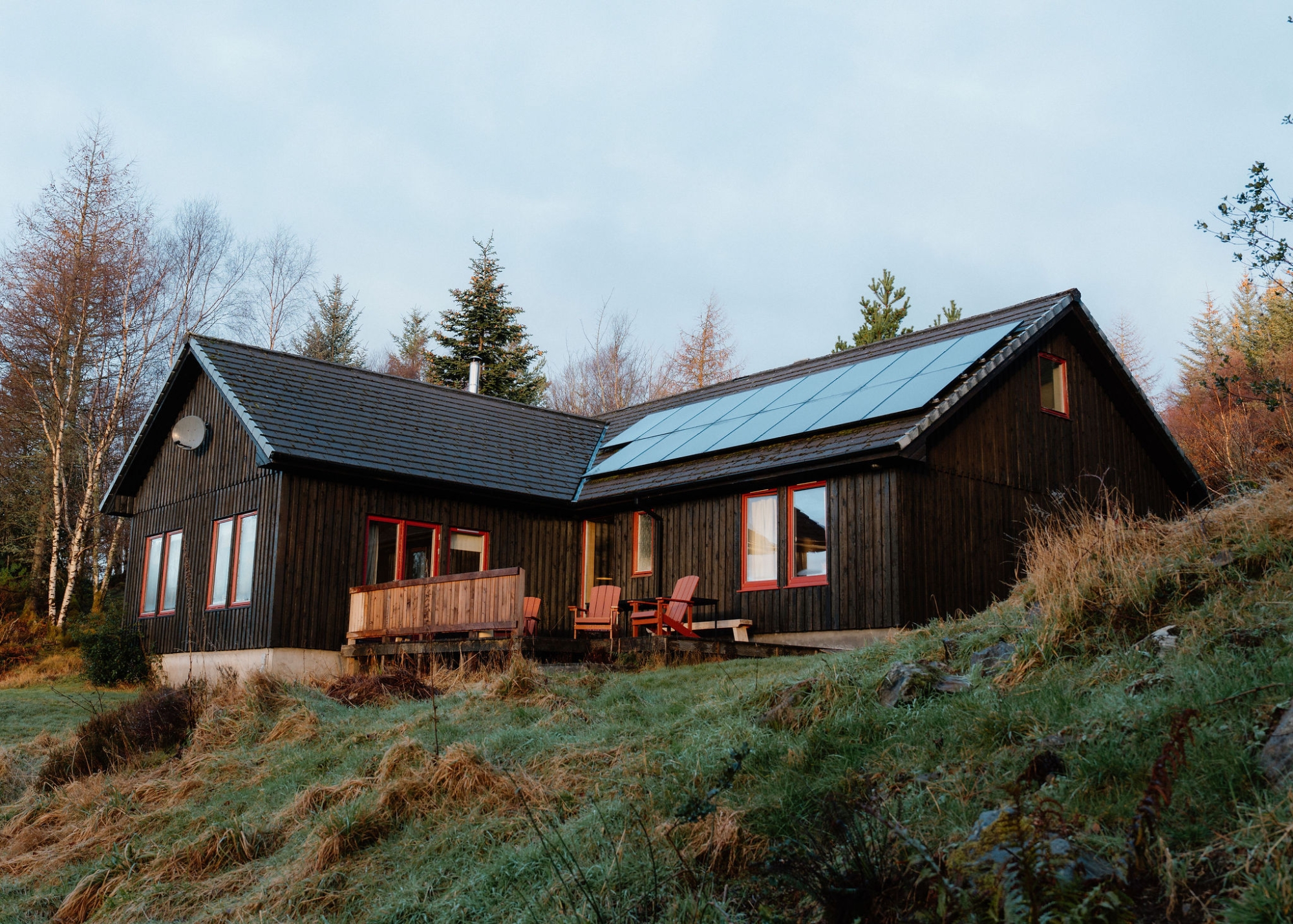 The house's gabled wing with a decked terrace and two chairs, solar panels along the roof, seen across a grassy slope.