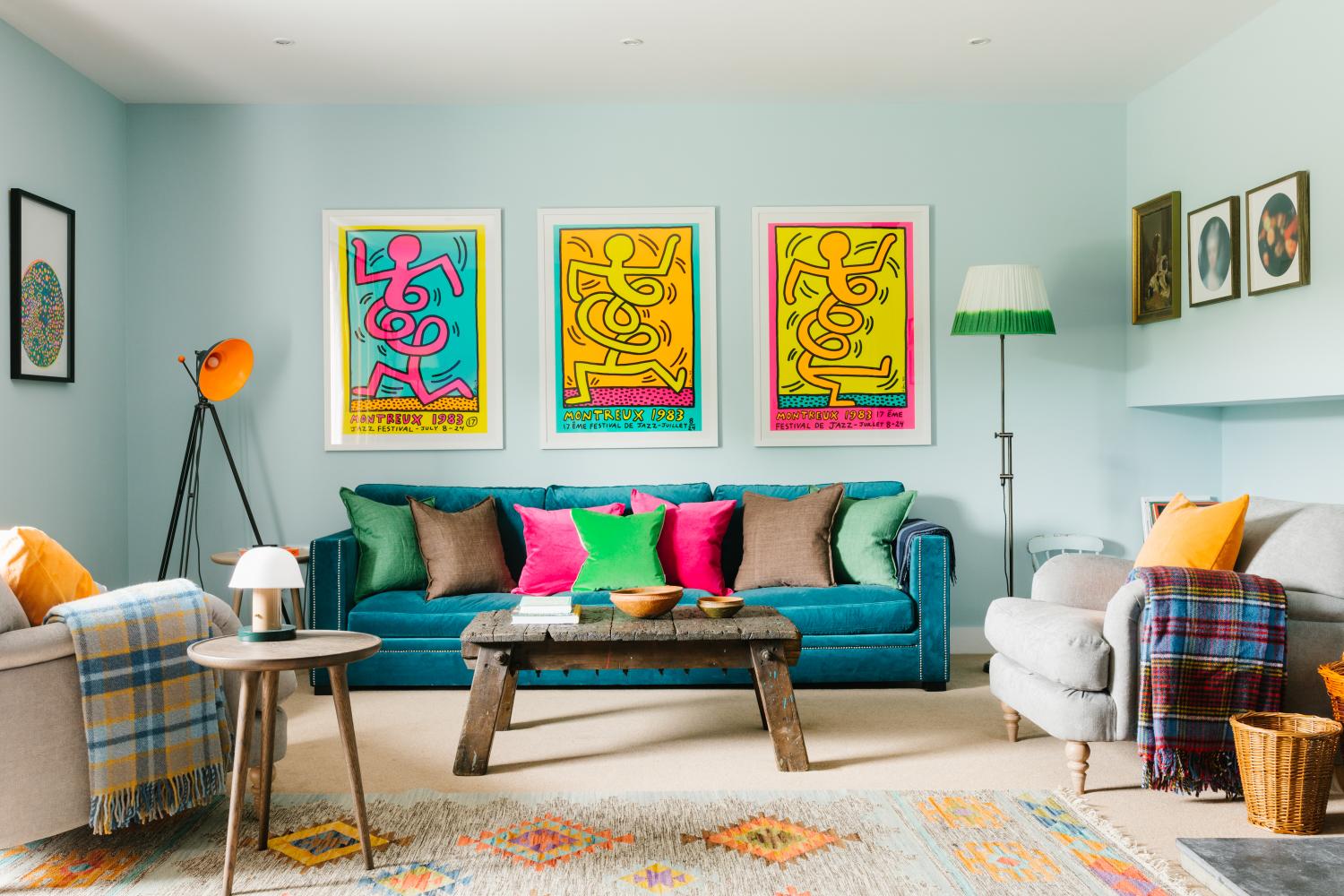 A teal velvet sofa with pink, green and brown cushions sits below three colourful pop-art prints, armchairs and a patterned rug completing the room.