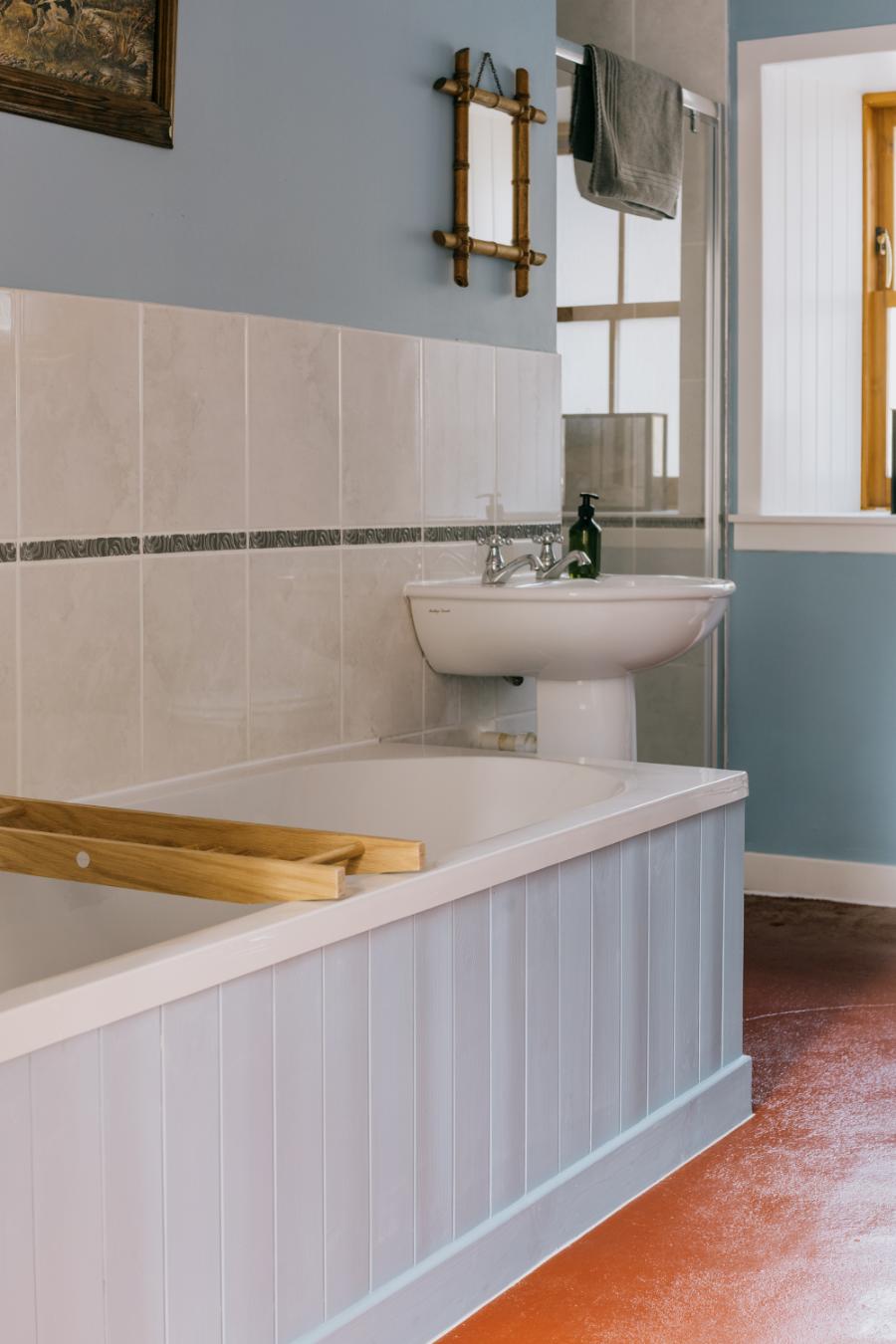 A pedestal basin stands beside a bath with a wooden bridge tray, a mirror and shower cubicle reflected against a pale blue wall.