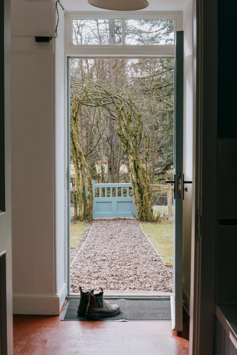 A pair of walking boots sits on the mat before an open front door, a mossy archway framing the gravel path beyond.