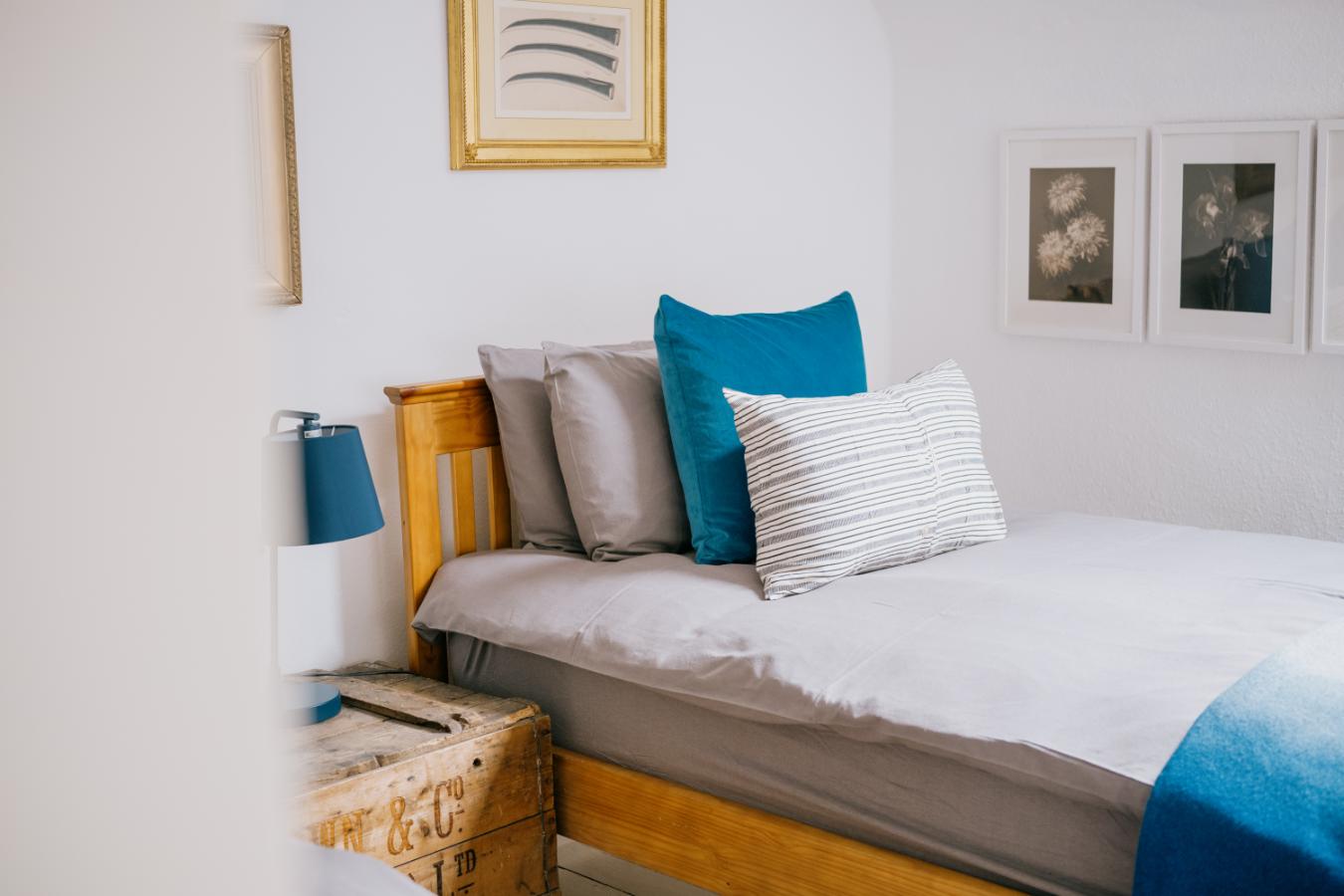 A bed with grey and teal cushions sits beneath framed prints, a wooden crate serving as a bedside table to the left.