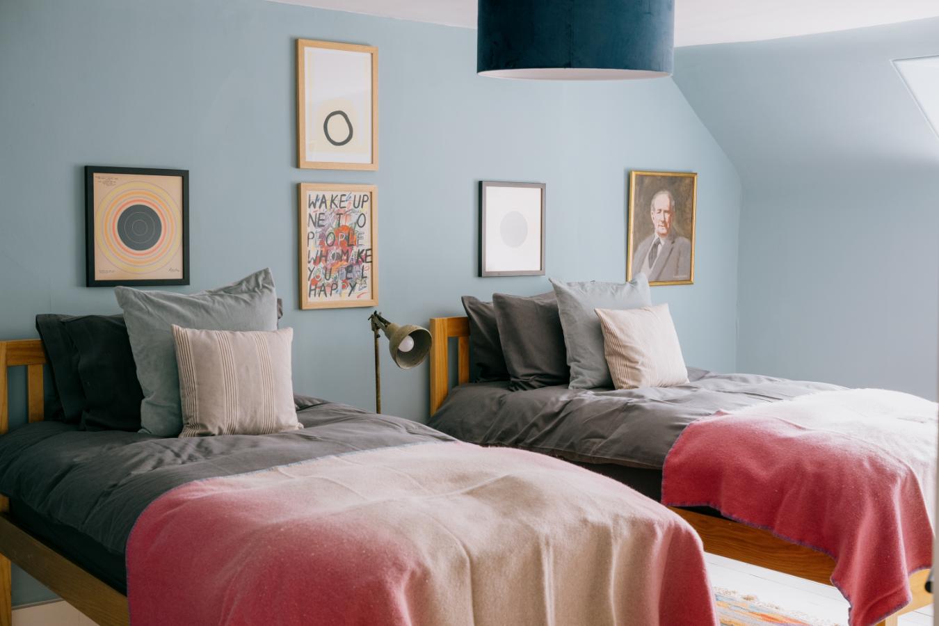 Twin beds with grey linen and pink throws sit beneath a gallery of framed prints, a brass lamp on the nightstand between them.