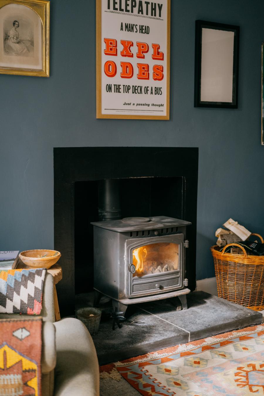 A wood burning stove is alight beside a wicker log basket, a framed print reading "Telepathy: A Man's Head Explodes on the Top Deck of a Bus" on the wall above.