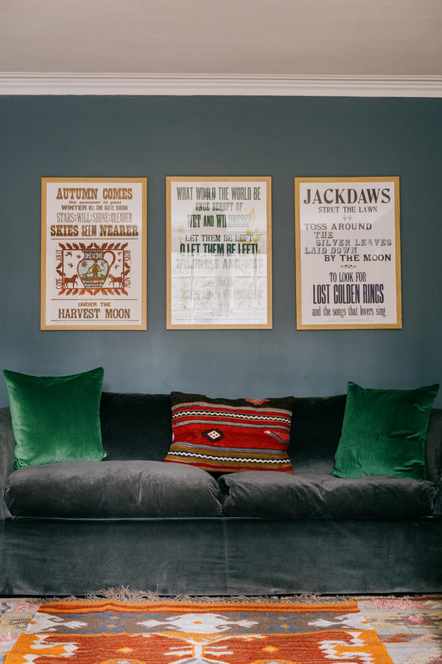 A green velvet sofa with a red patterned cushion sits below three framed poems including "Autumn Comes" and "Jackdaws", on a patterned rug.