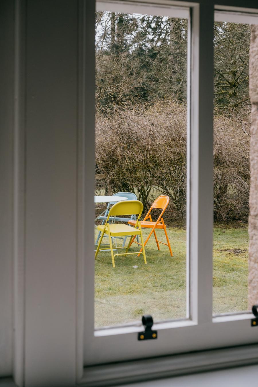 Yellow and orange folding chairs stand around a table on a lawn edged by bare hedging, seen through a window.