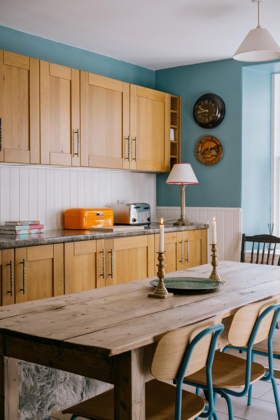 A wooden dining table with teal chairs stands before an antique clock and plate mounted on a teal wall, an orange bread bin on the counter behind.