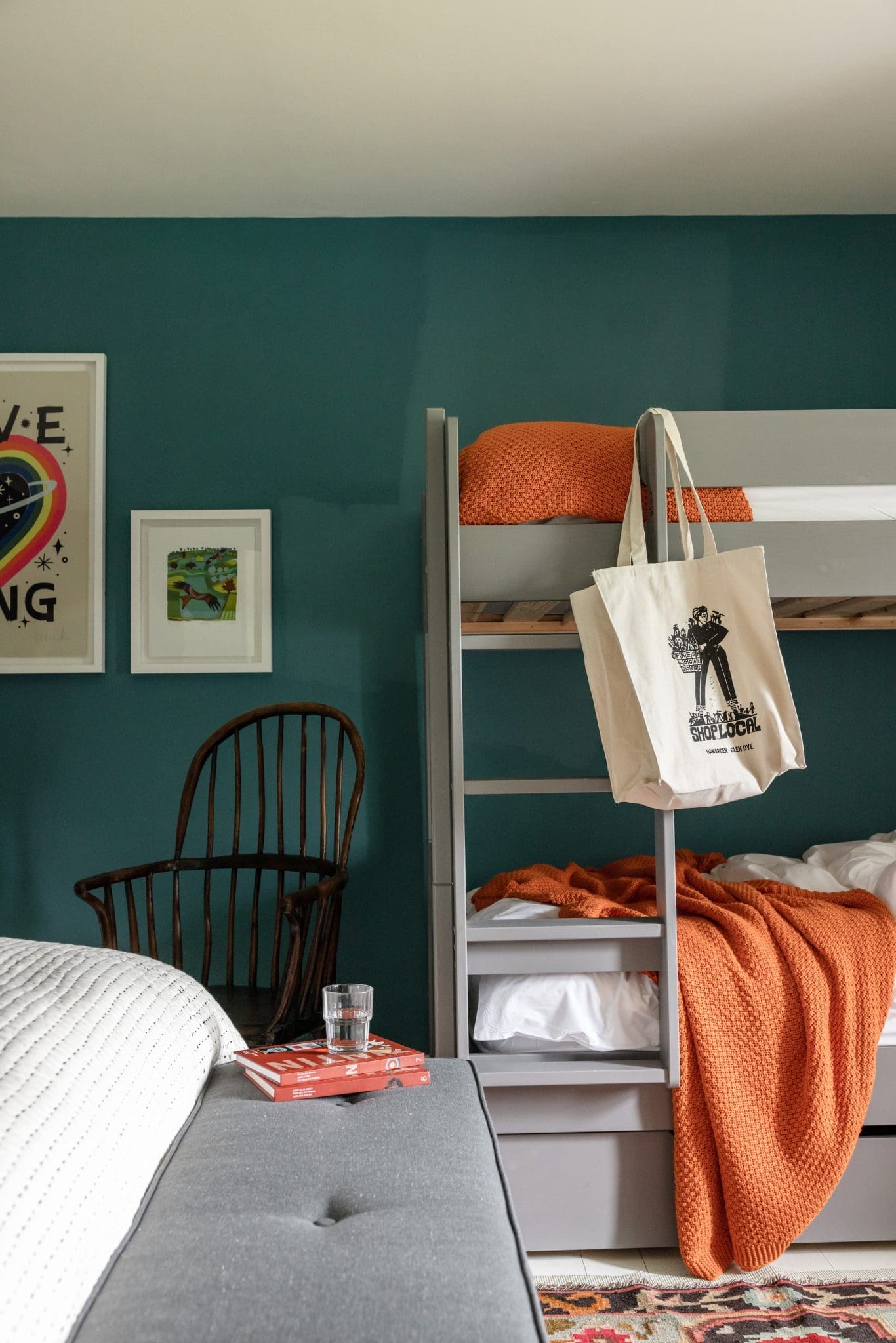 A grey bunk bed with an orange throw stands against a dark green wall, a canvas tote bag hanging from the ladder and a windsor chair with framed prints to the side.