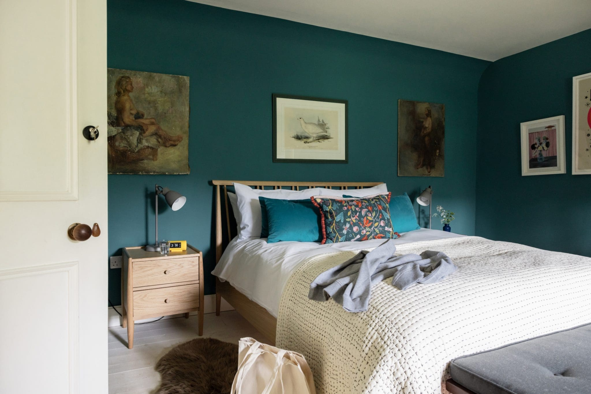 A bed with patterned cushions sits before a teal wall hung with framed paintings, a wooden nightstand with lamps to the left.