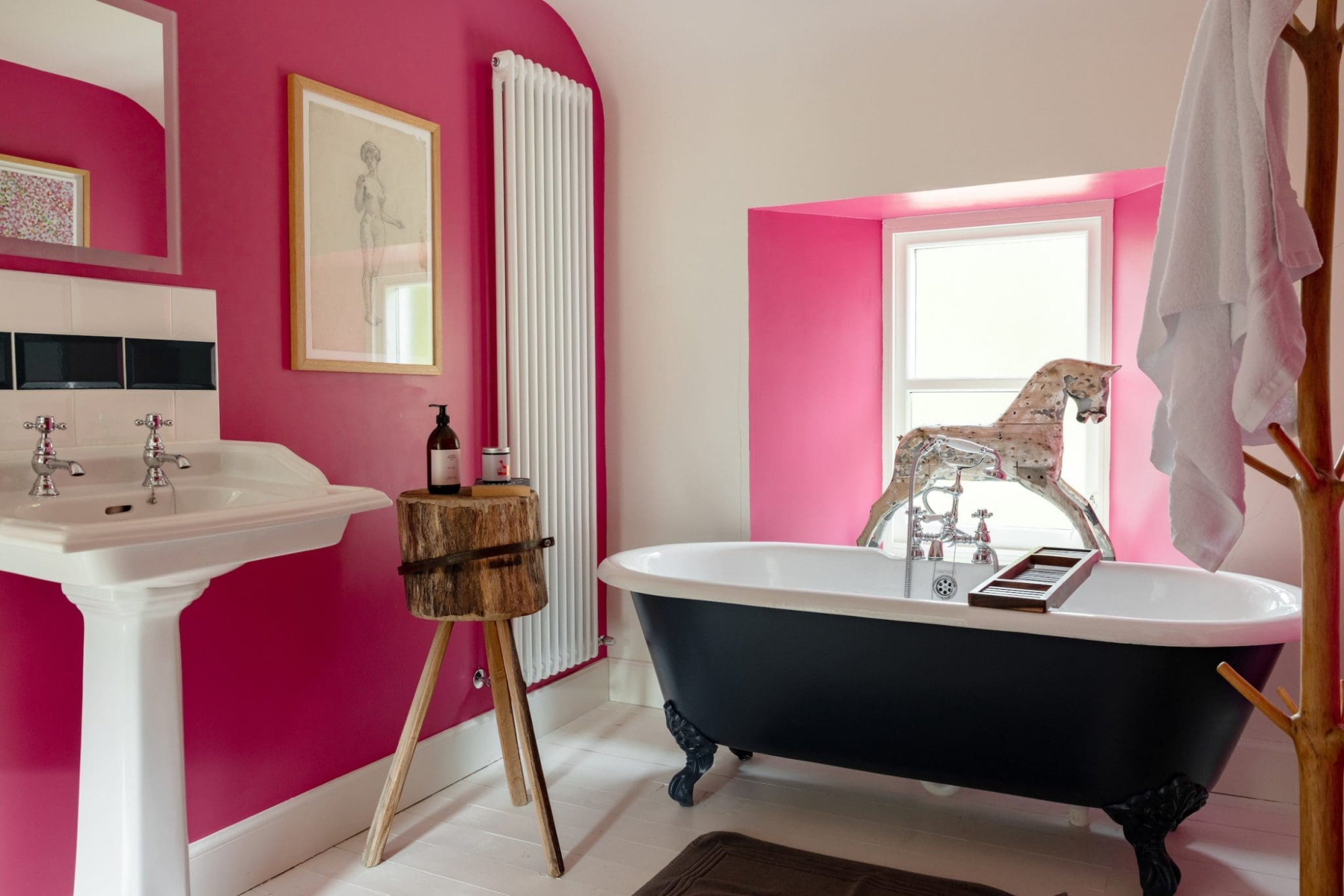A black roll-top bath with a horse-shaped tap stands before a pink alcove window, a pedestal basin and wooden stool to the left.