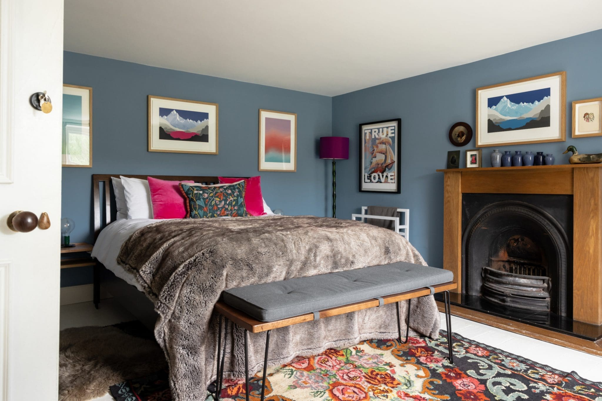 A bed with pink and floral cushions sits before framed art including a print reading "True Love", an open fireplace and patterned rug completing the room.
