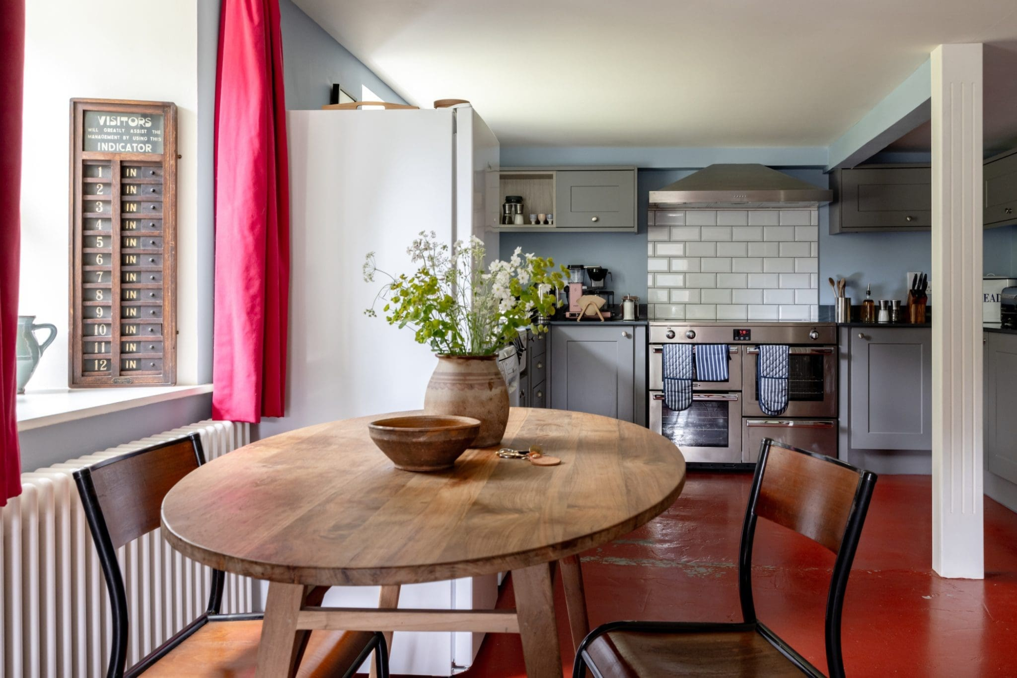 A round wooden table with two dark chairs sits beneath a red curtain, a stoneware jug of flowers at its centre, with a grey fitted kitchen and white tiled splashback behind and a dark red painted floor underfoot.