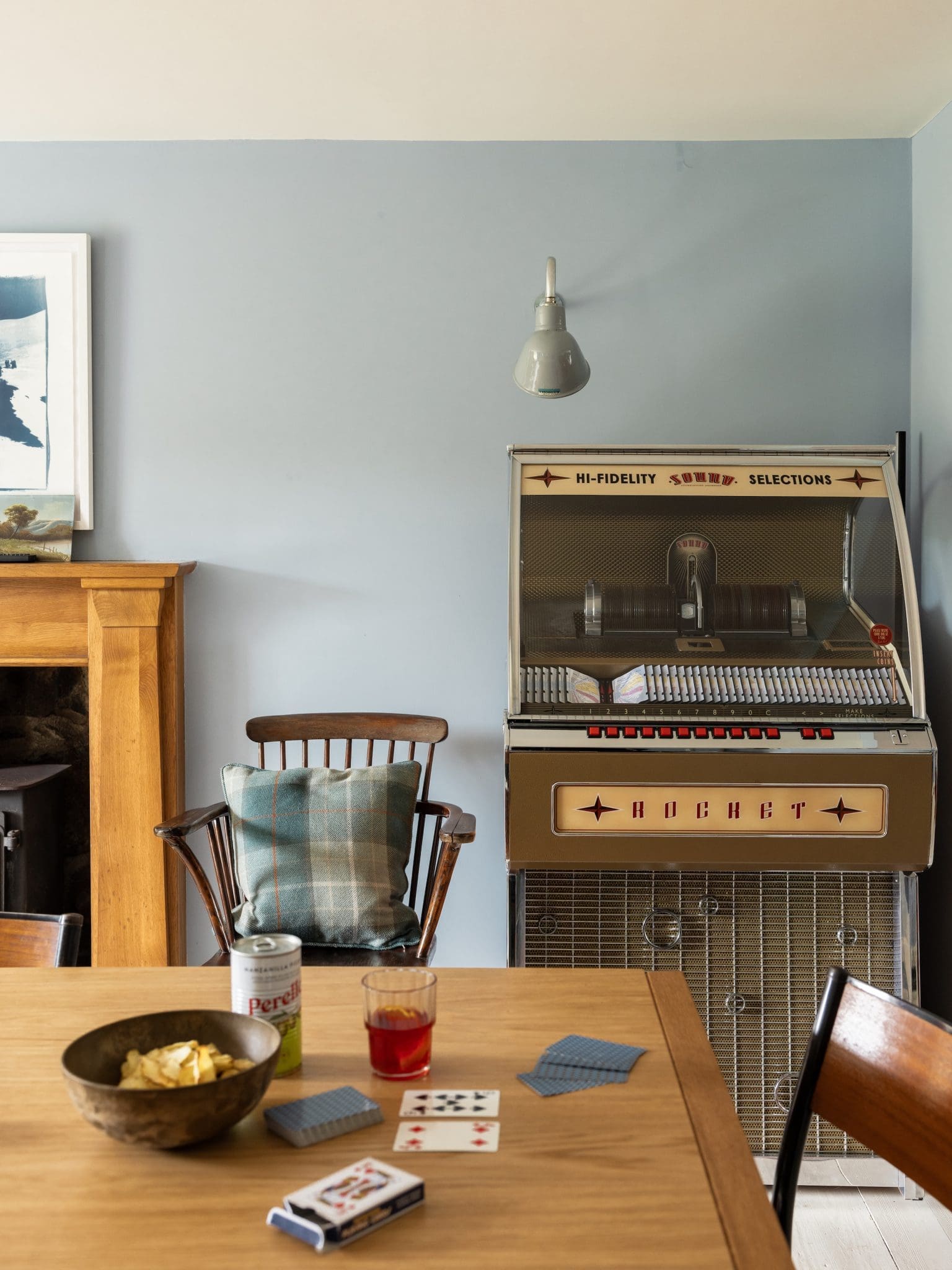A vintage jukebox stands in the corner of a pale blue room beside a wall-mounted lamp and a wooden armchair with a checked cushion, with playing cards and a bowl of crisps set on a table in the foreground.