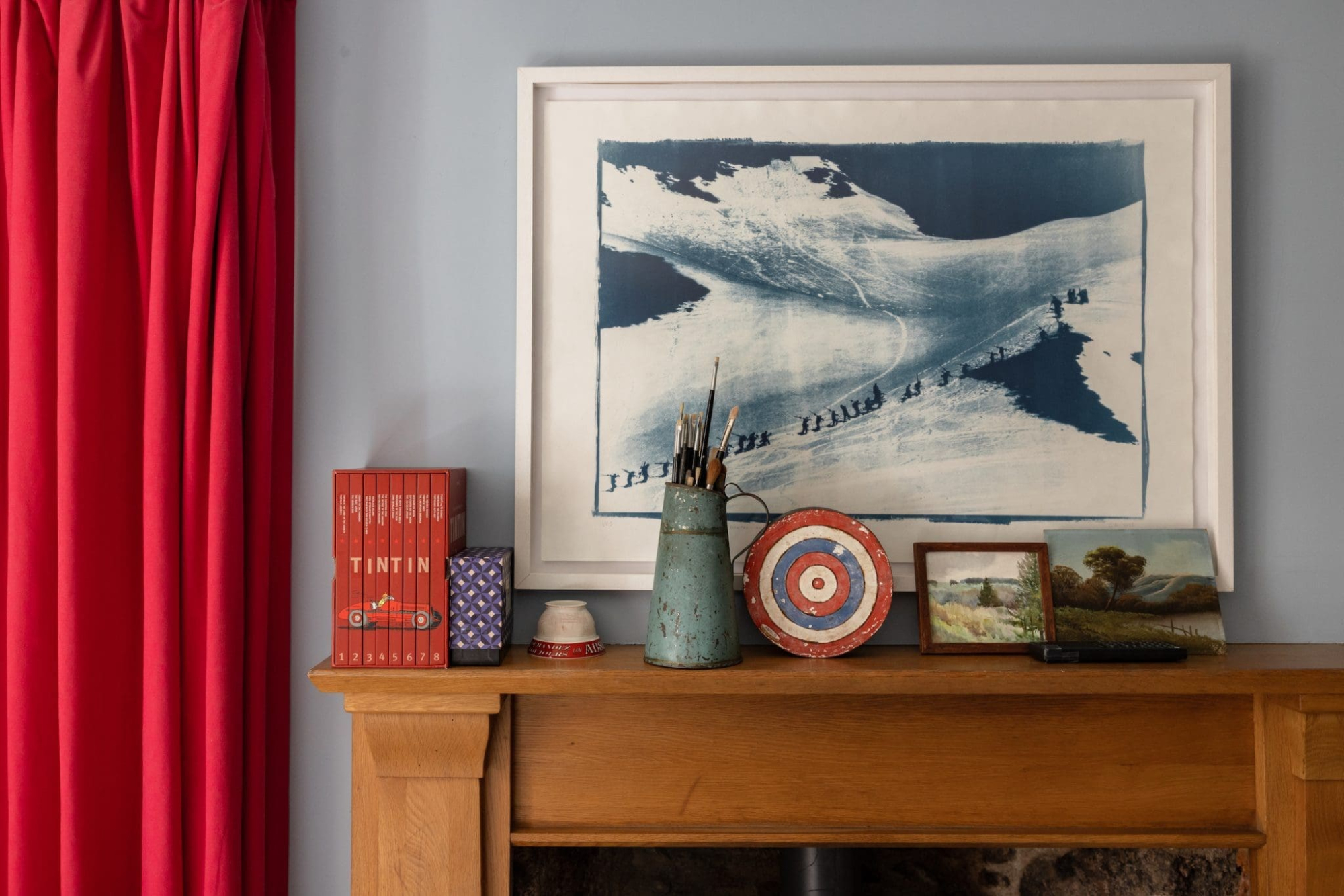 A wooden fireplace mantelpiece holds a pot of paintbrushes, a target-patterned dish, small framed pictures and a boxed set of "Tintin" books, with a framed cyanotype landscape print above and a red curtain to the left.