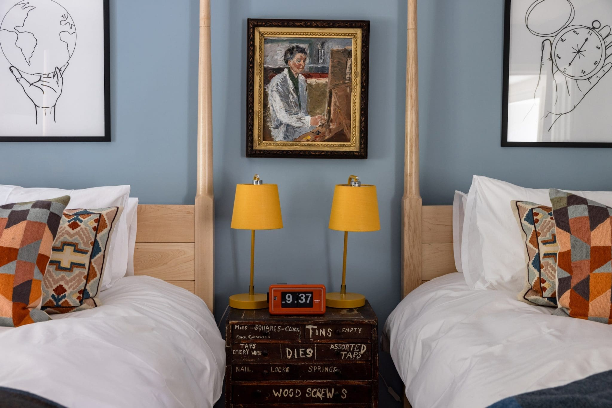 Twin beds with patterned cushions flank a vintage stacked crate nightstand holding two yellow lamps and a clock, a framed portrait above.