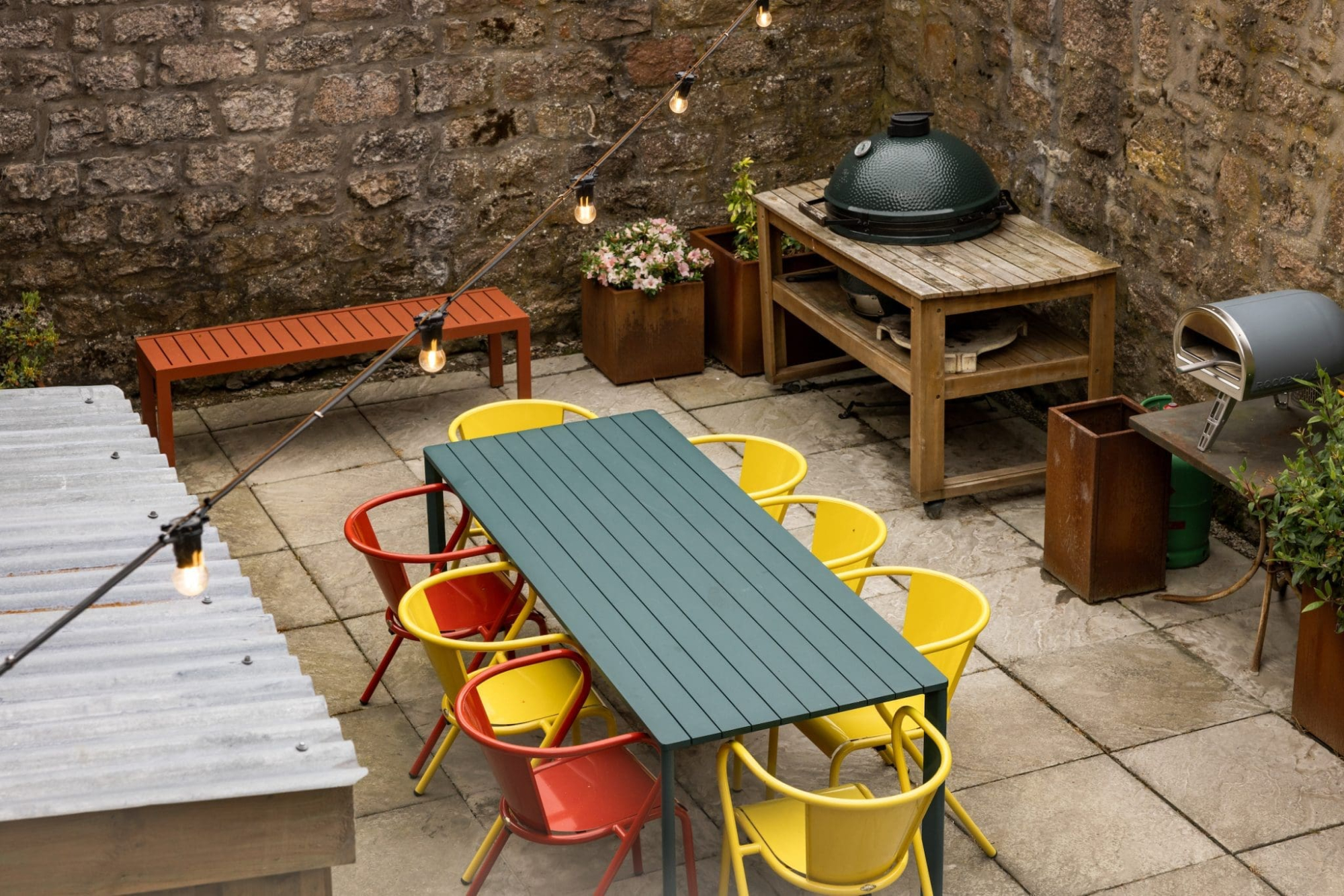 A paved courtyard garden has a long dark green table surrounded by yellow and orange chairs, string lights overhead, a domed barbecue on a timber stand, and potted plants along a stone wall.