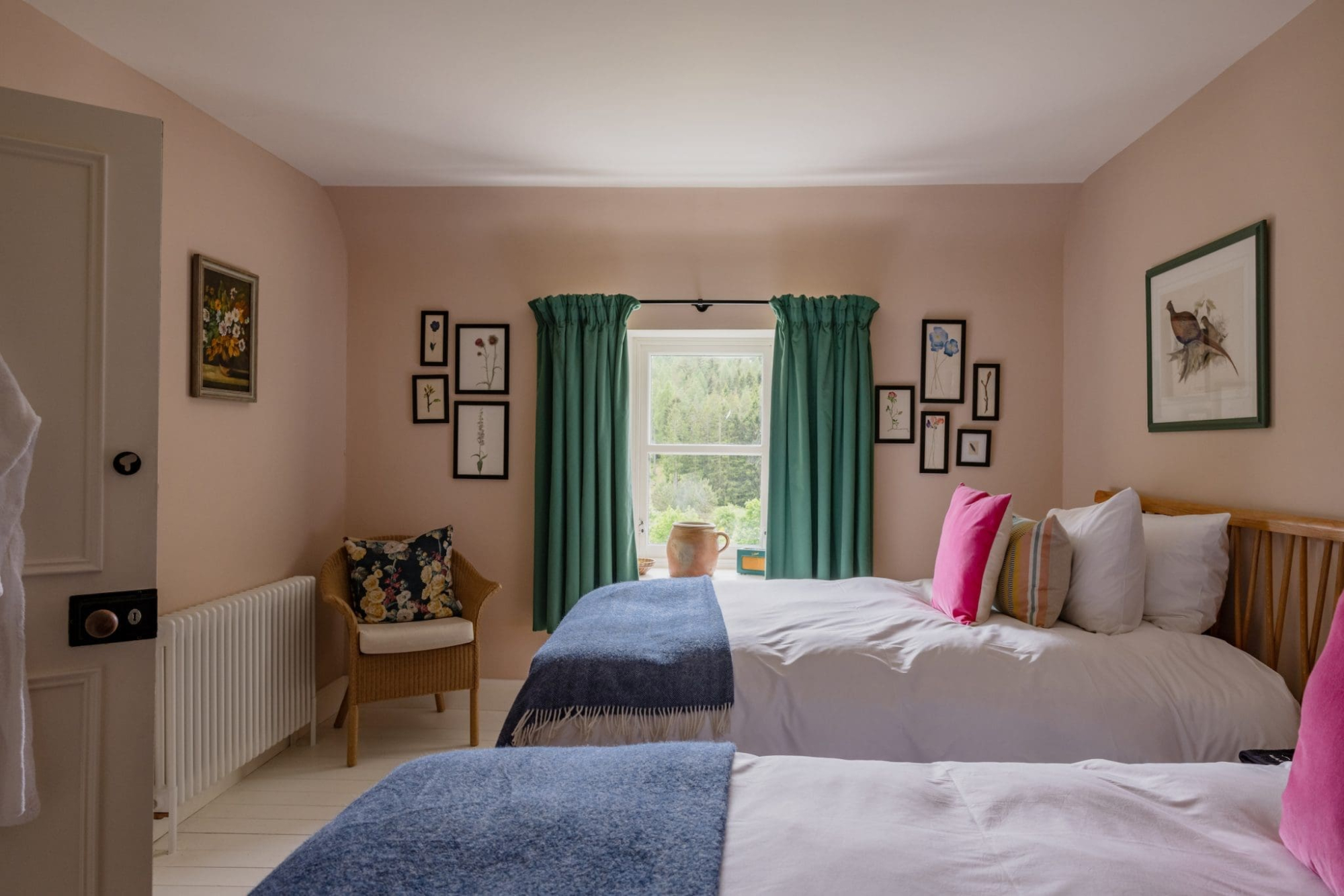 Twin beds with pink cushions sit before a curtained window, framed prints lining the pink walls and a wicker chair in the corner.