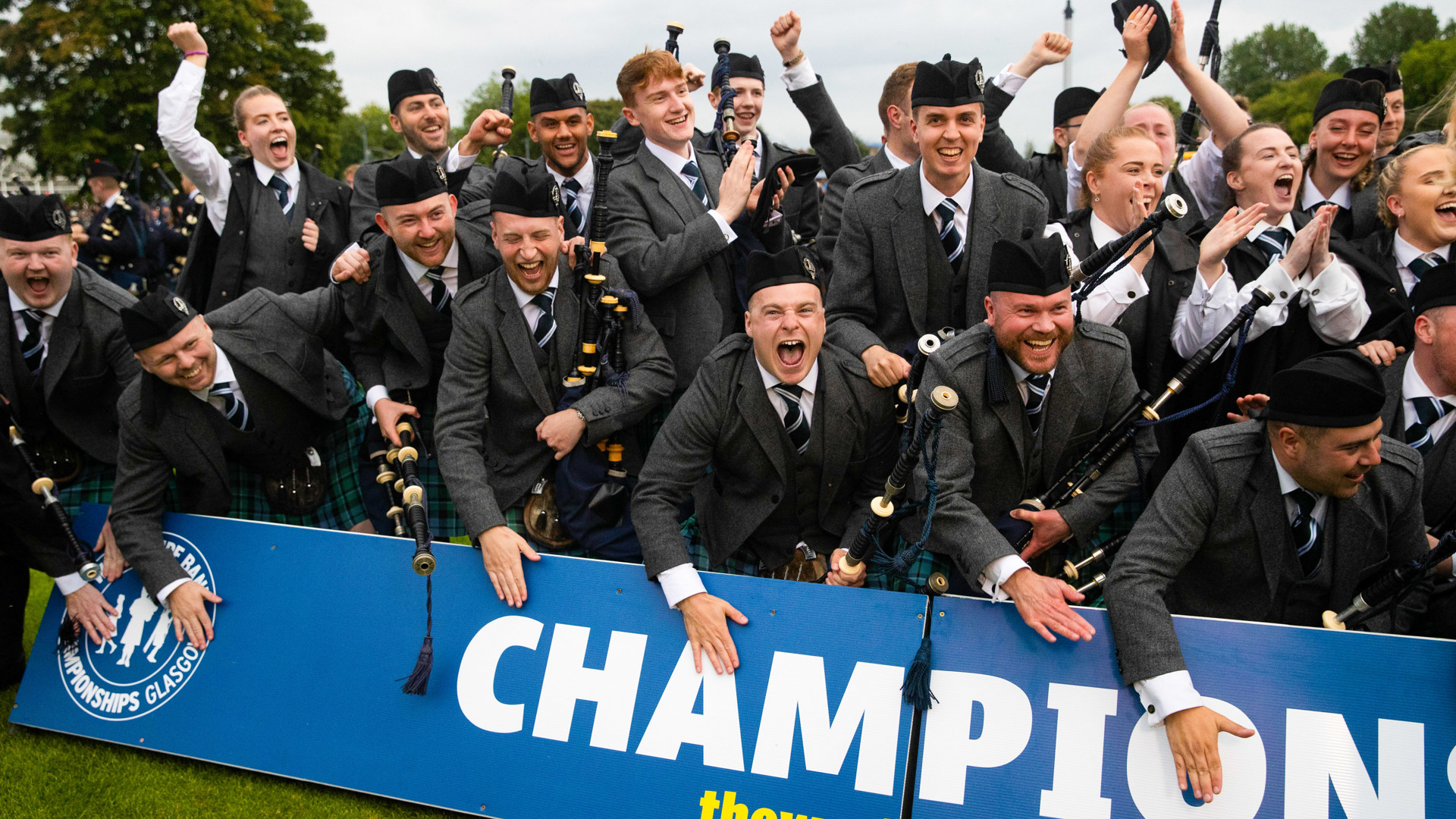 Pipe band members cheer behind a blue Champions barrier