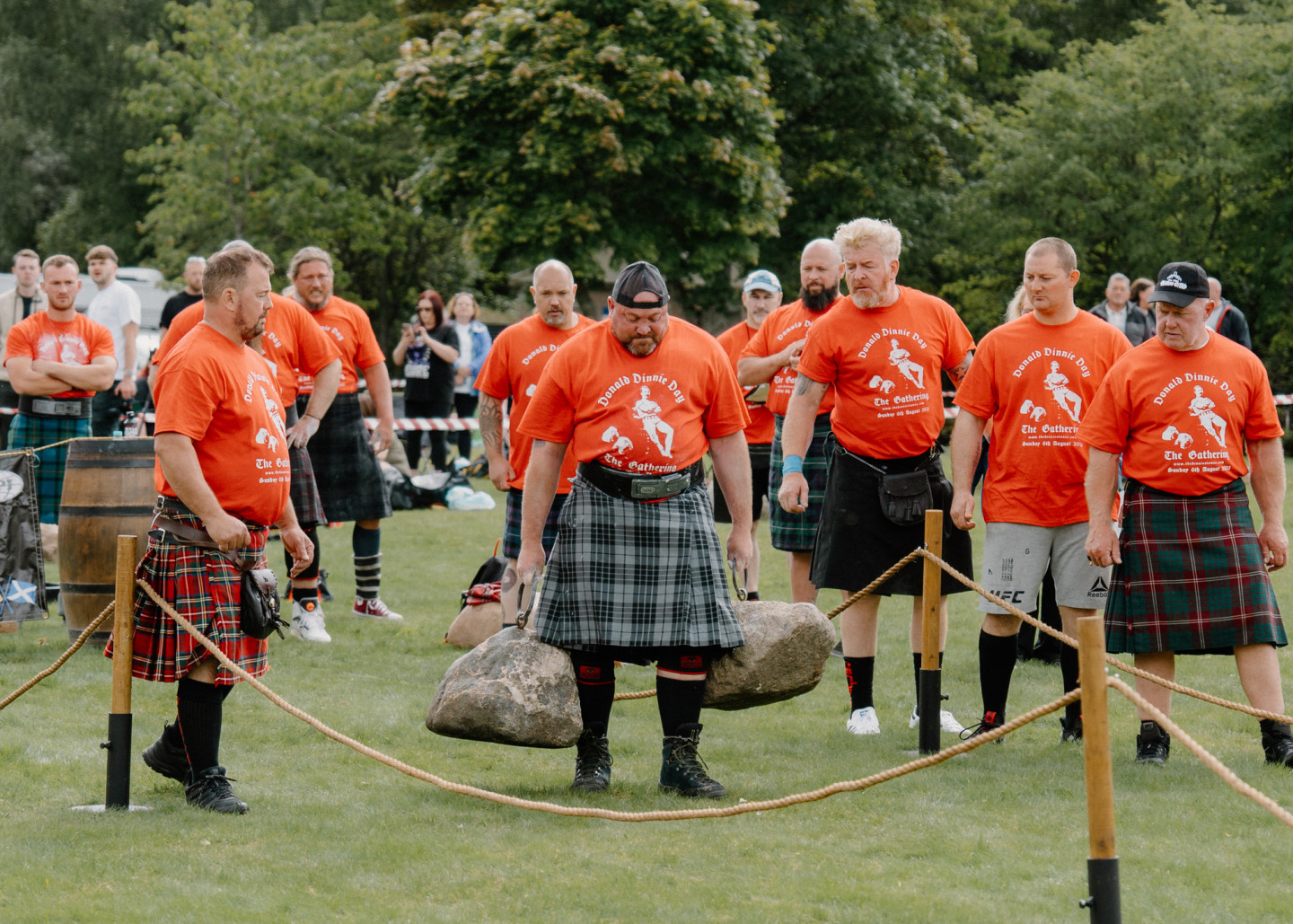 Kilted athletes in orange Highland Games T-shirts watch a stone-lifting competitor