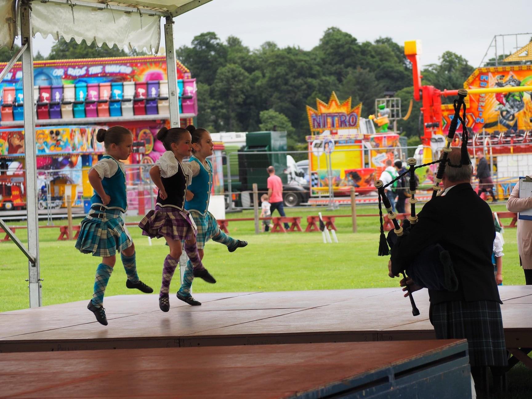 Highland dancers leap on stage as a piper plays beside the Nitro fairground ride