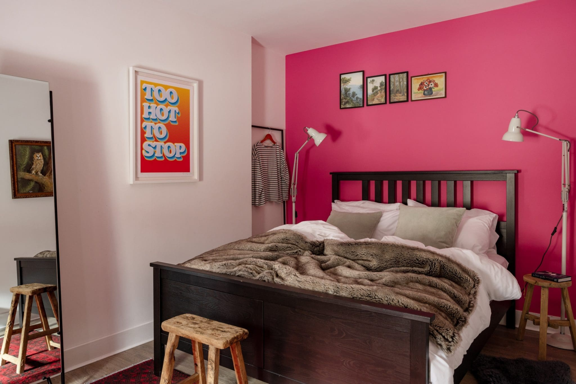A dark wood bed with a fur throw stands against a bright pink wall hung with small framed pictures and a print reading "TOO HOT TO STOP", a gilt-framed portrait reflected in a mirror to the side.