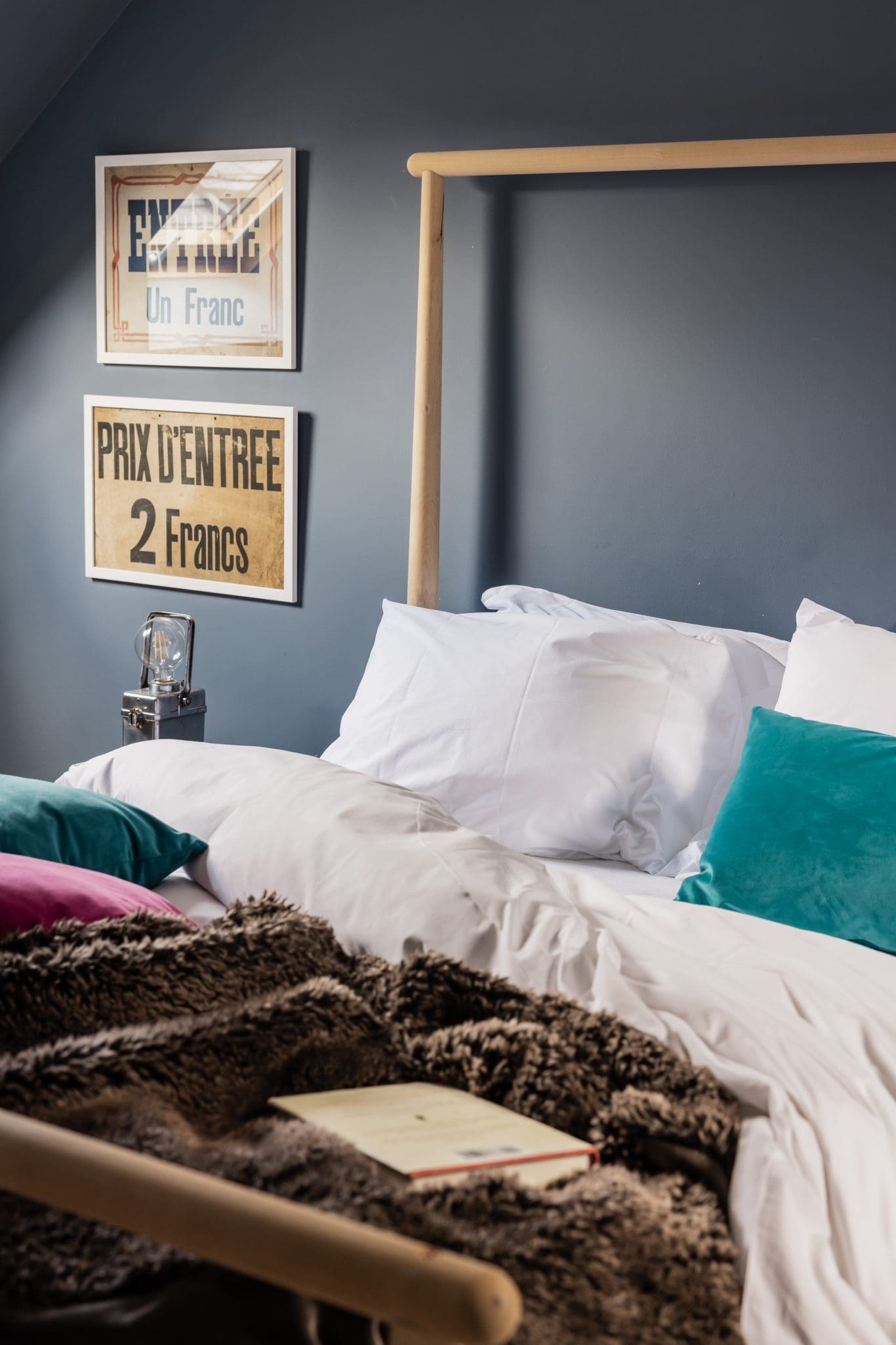 A wooden bed with white pillows and a fur throw stands beneath two framed vintage prints reading "ENTREE Un Franc" and "PRIX D'ENTREE 2 Francs" on a dark blue wall.
