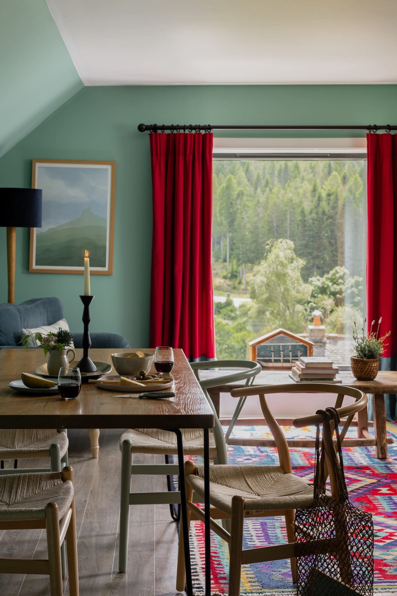 A wooden dining table set with wine glasses and a lit candle sits before a large window framed by red curtains, dense trees visible outside and a rug of patterned kilim underfoot.