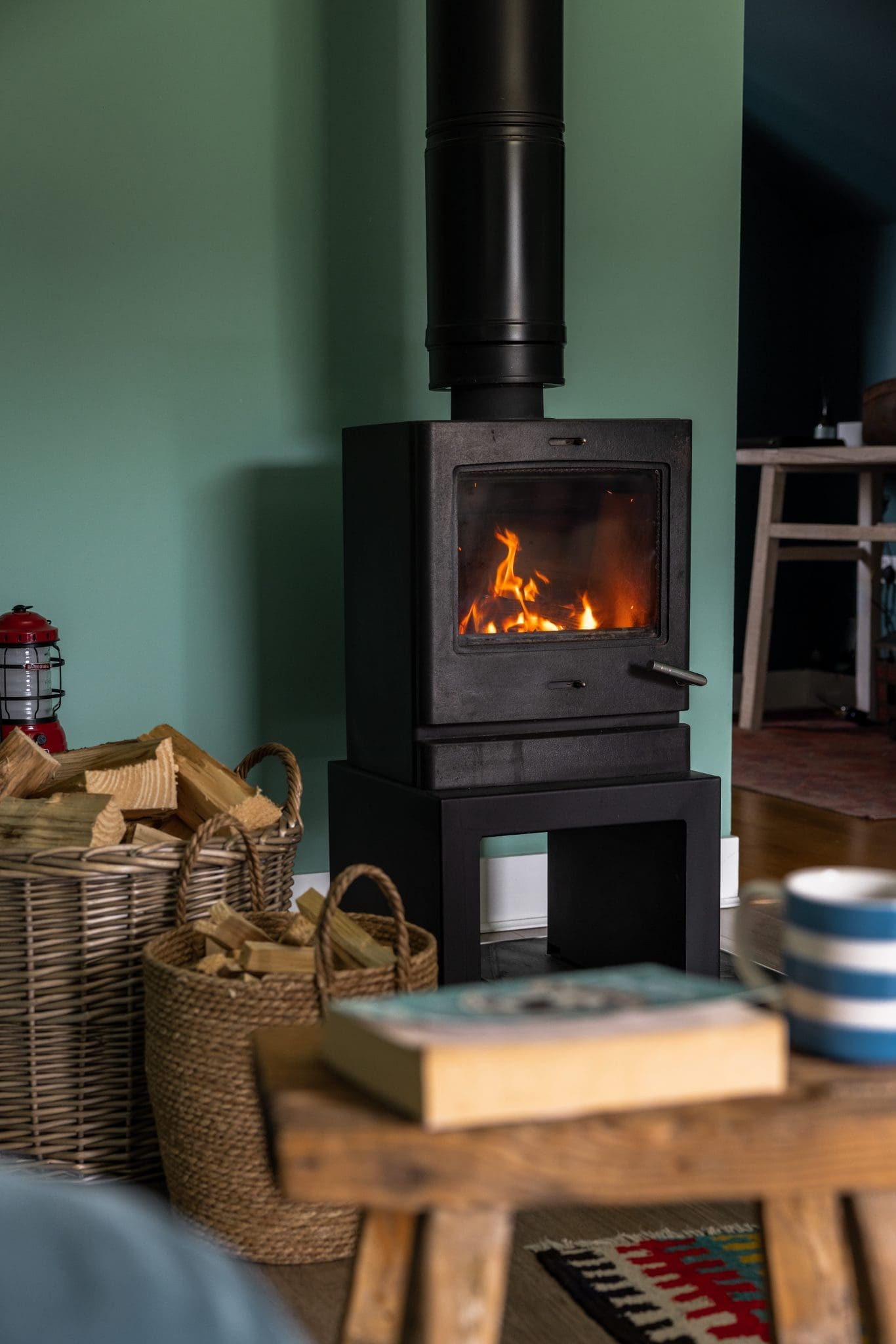 A black wood-burning stove is lit between two baskets of logs, a wooden bench in the foreground holding a book and a striped mug.