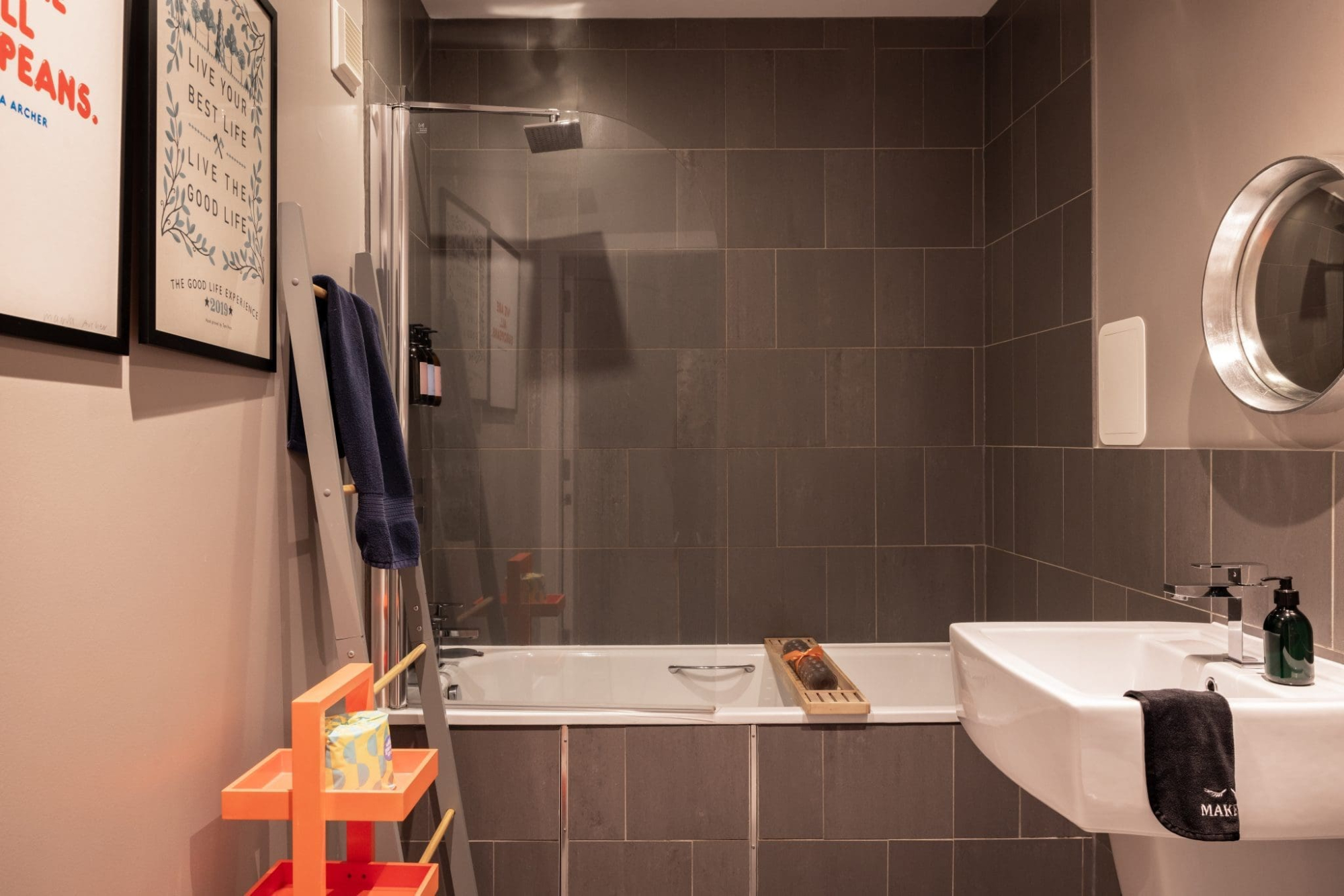 A glass shower screen stands over a bath in a grey-tiled bathroom, a round mirror above the sink and an orange shelving unit of toiletries in the corner.