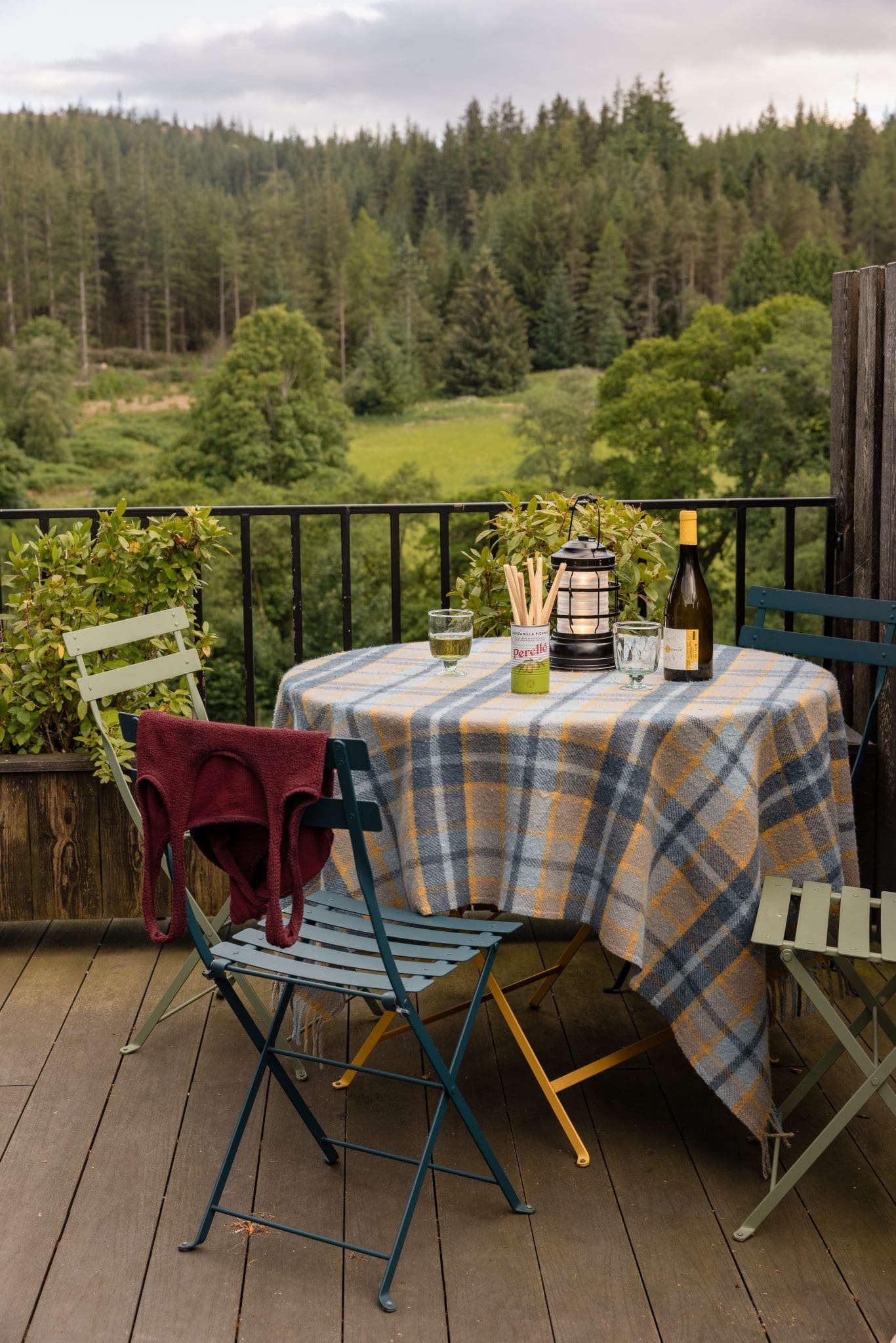 A round table laid with a plaid cloth holds glasses, a lit lantern and a bottle of wine, folding chairs around it and forest visible beyond the railing.