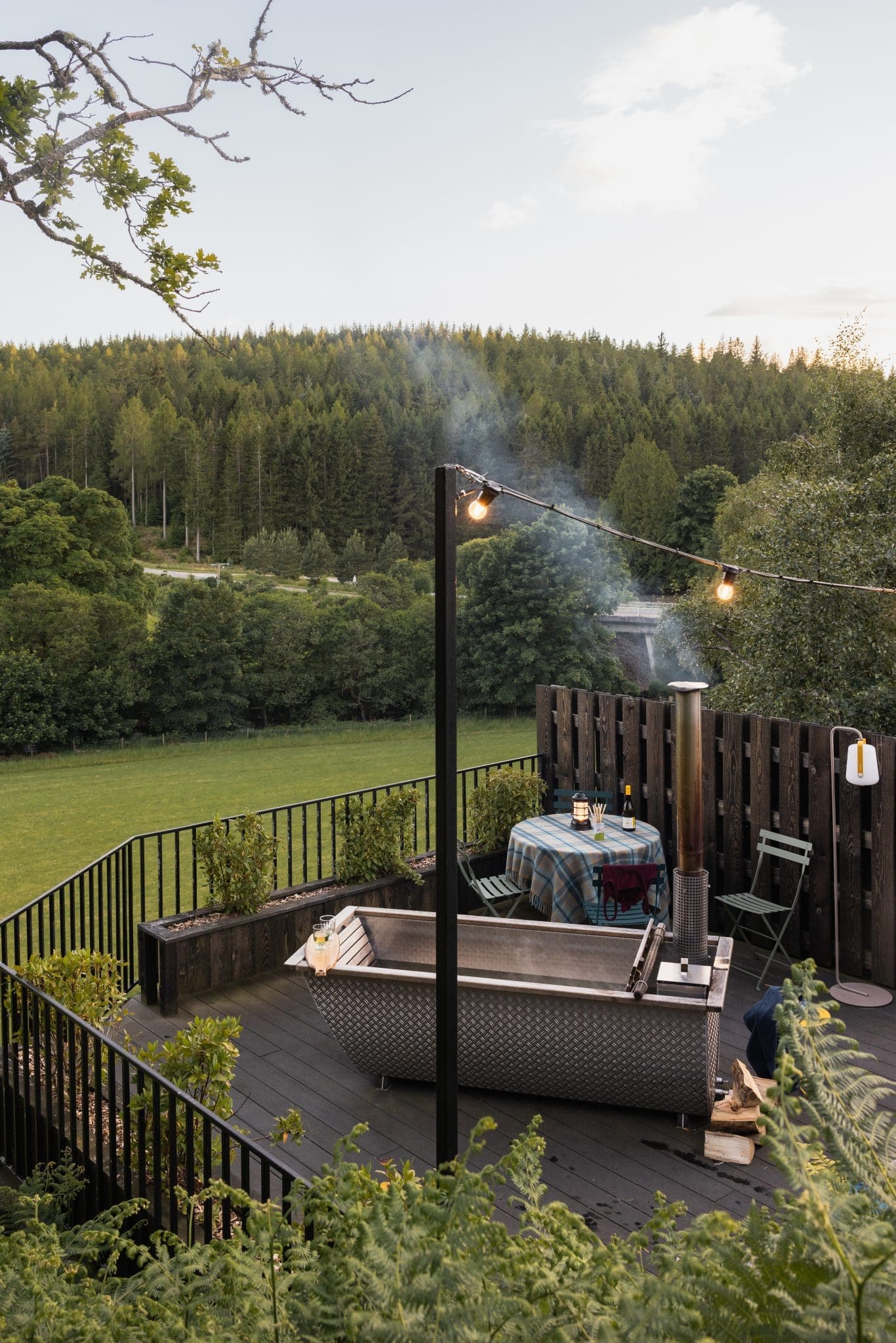 A wood-fired hot tub sends smoke into the evening air on a deck strung with festoon lights, a curved rusted roof and trees framing the scene.