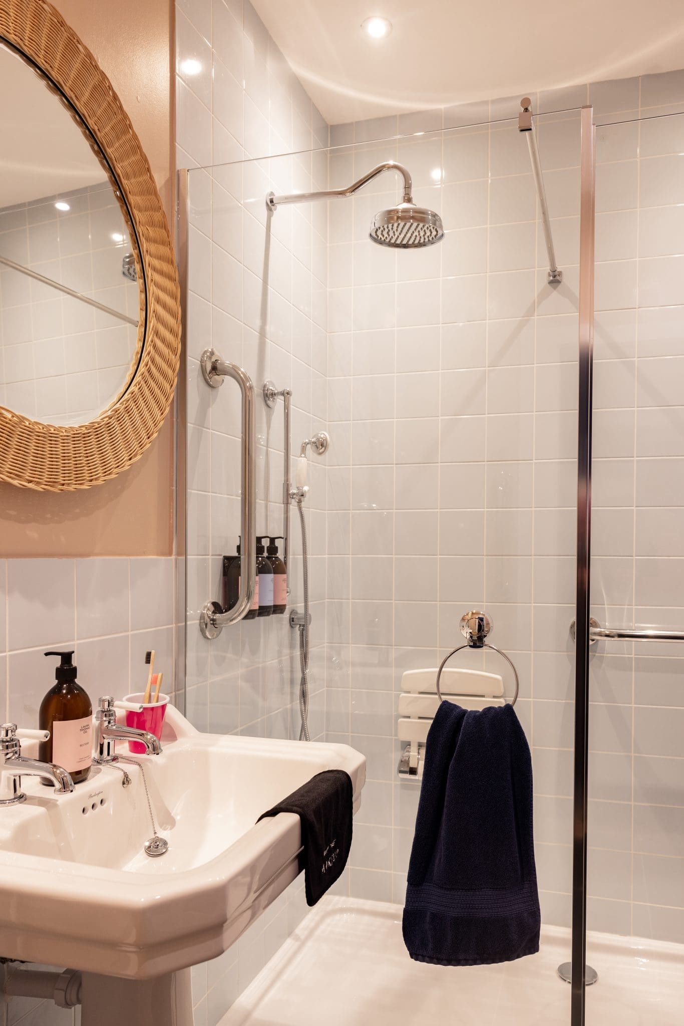 A walk-in shower with a rainfall head and grab rail stands beside a sink with soap dispensers and navy towels, a round rattan-framed mirror on the wall.