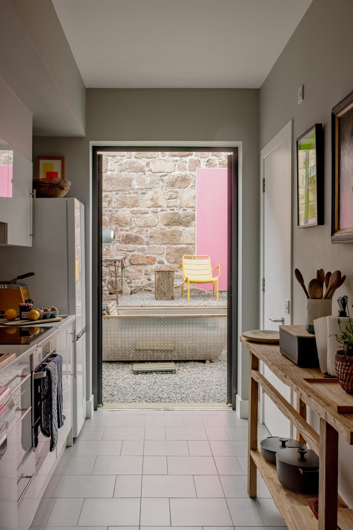 A galley kitchen leads through an open door to a paved courtyard with a stone wall, a pink panel and a yellow chair, a console table of wooden spoons and pans on the right.