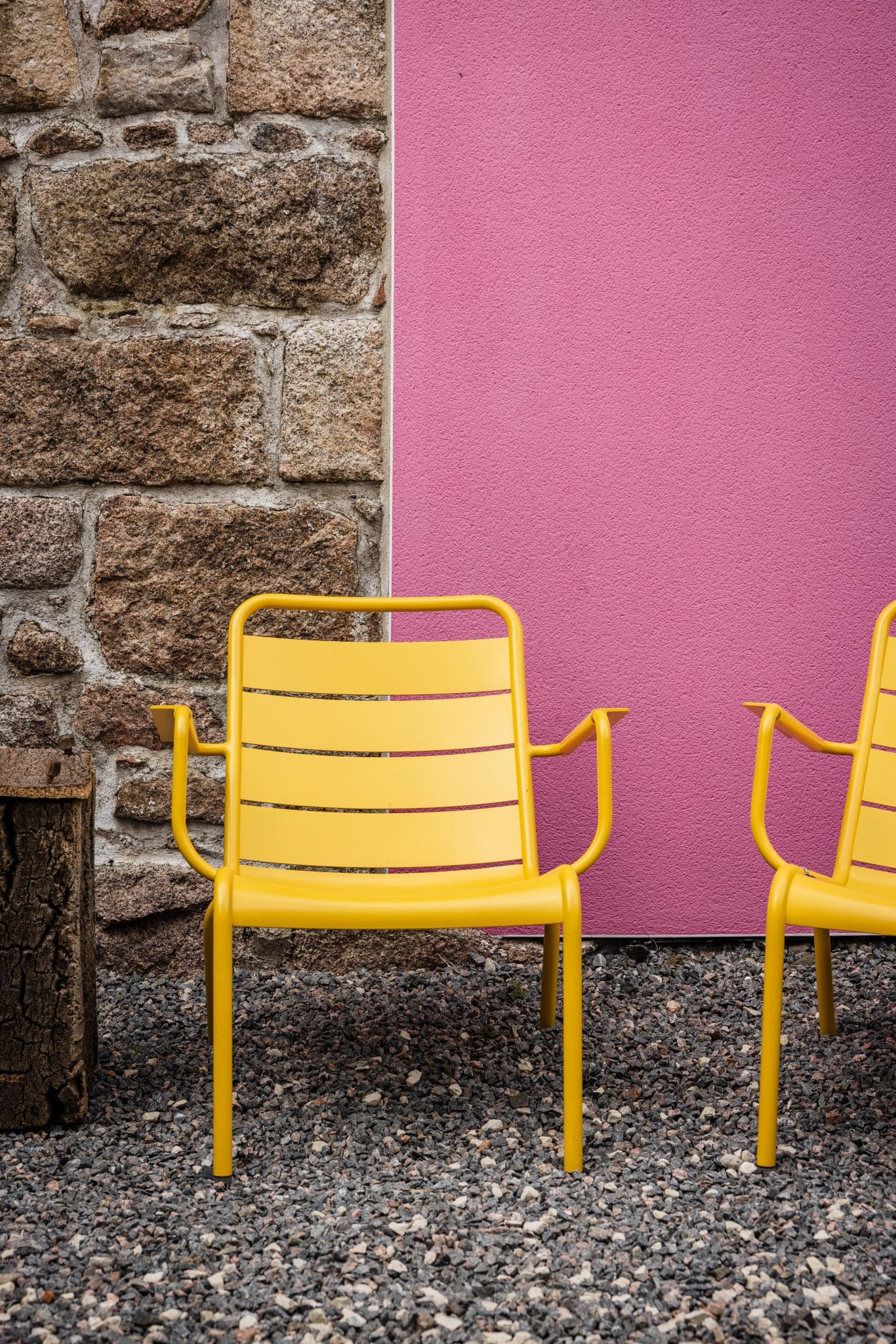 Two yellow metal armchairs with armrests stand on gravel against a stone wall and a pink painted panel.