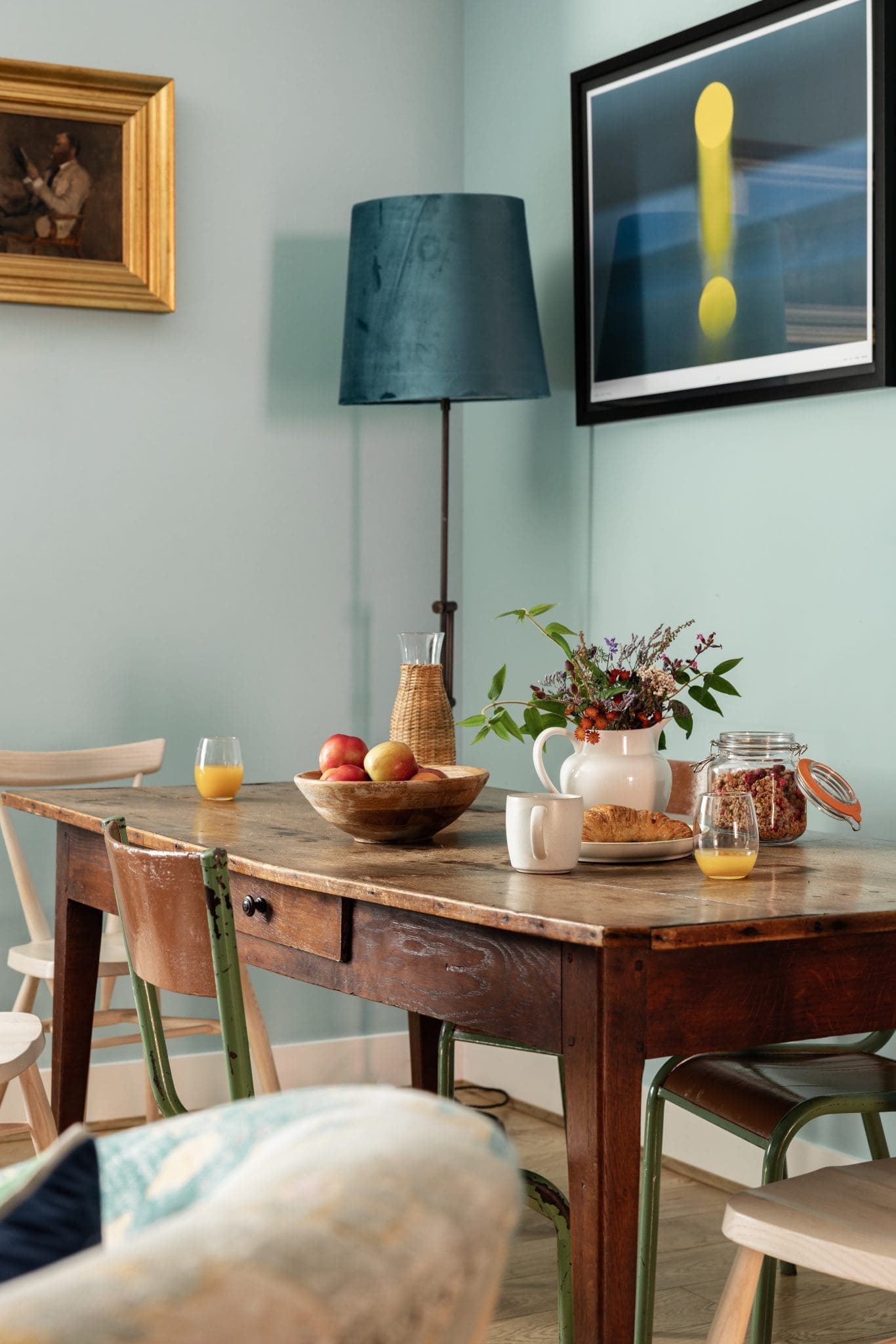 A wooden table set for breakfast with a fruit bowl, a jug of flowers, a jar of granola and glasses of orange juice sits below a teal-shaded floor lamp and a gilt-framed painting.