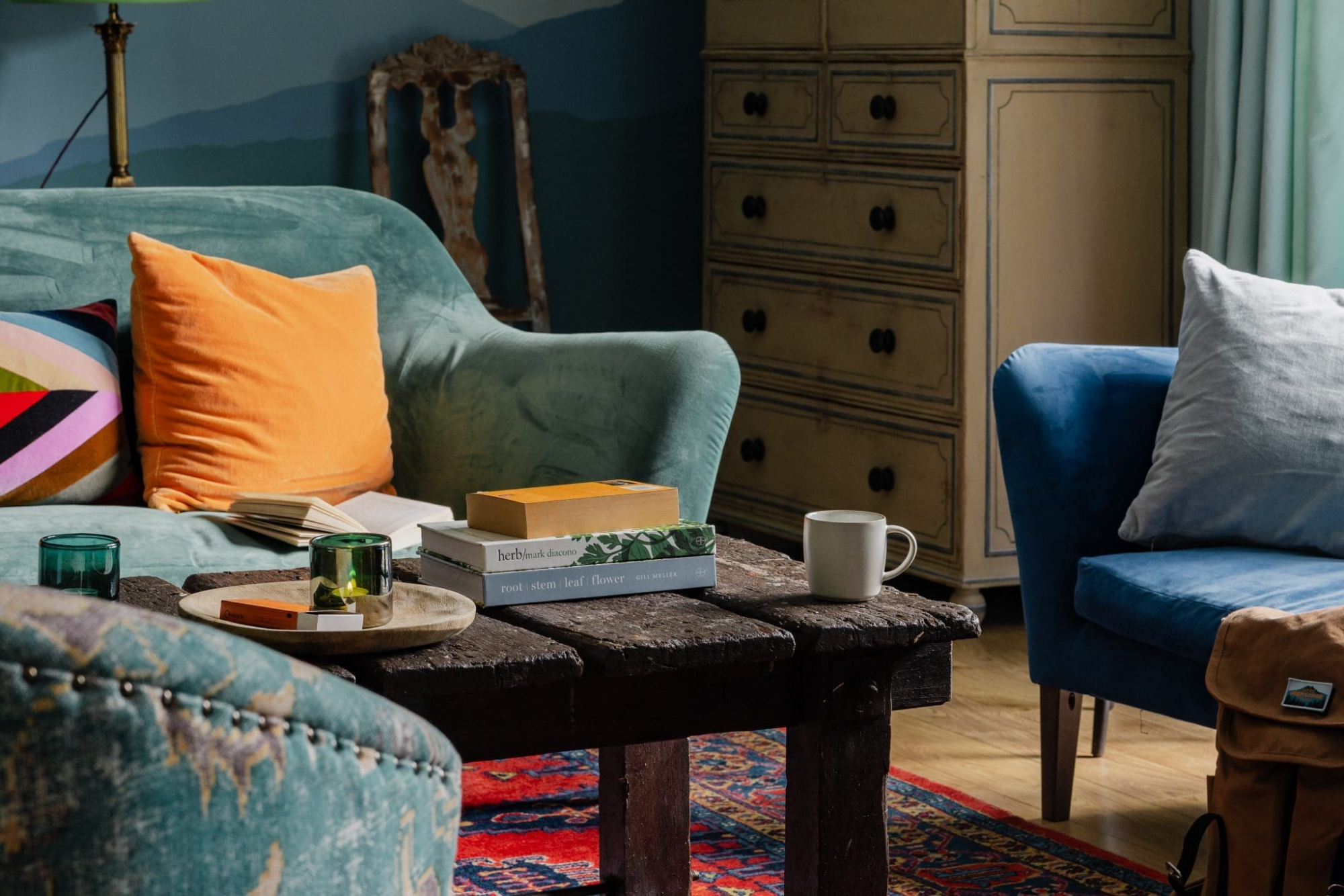 A teal sofa with an orange cushion sits beside a wooden trunk coffee table holding books and a mug, a dark wood chest of drawers and a blue armchair with a grey cushion nearby.