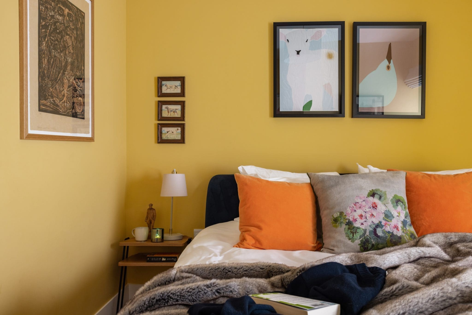 A mustard-yellow bedroom has a navy headboard with orange cushions and a floral cushion, a fur throw across the bed, and framed illustrations of an animal and a bird above.