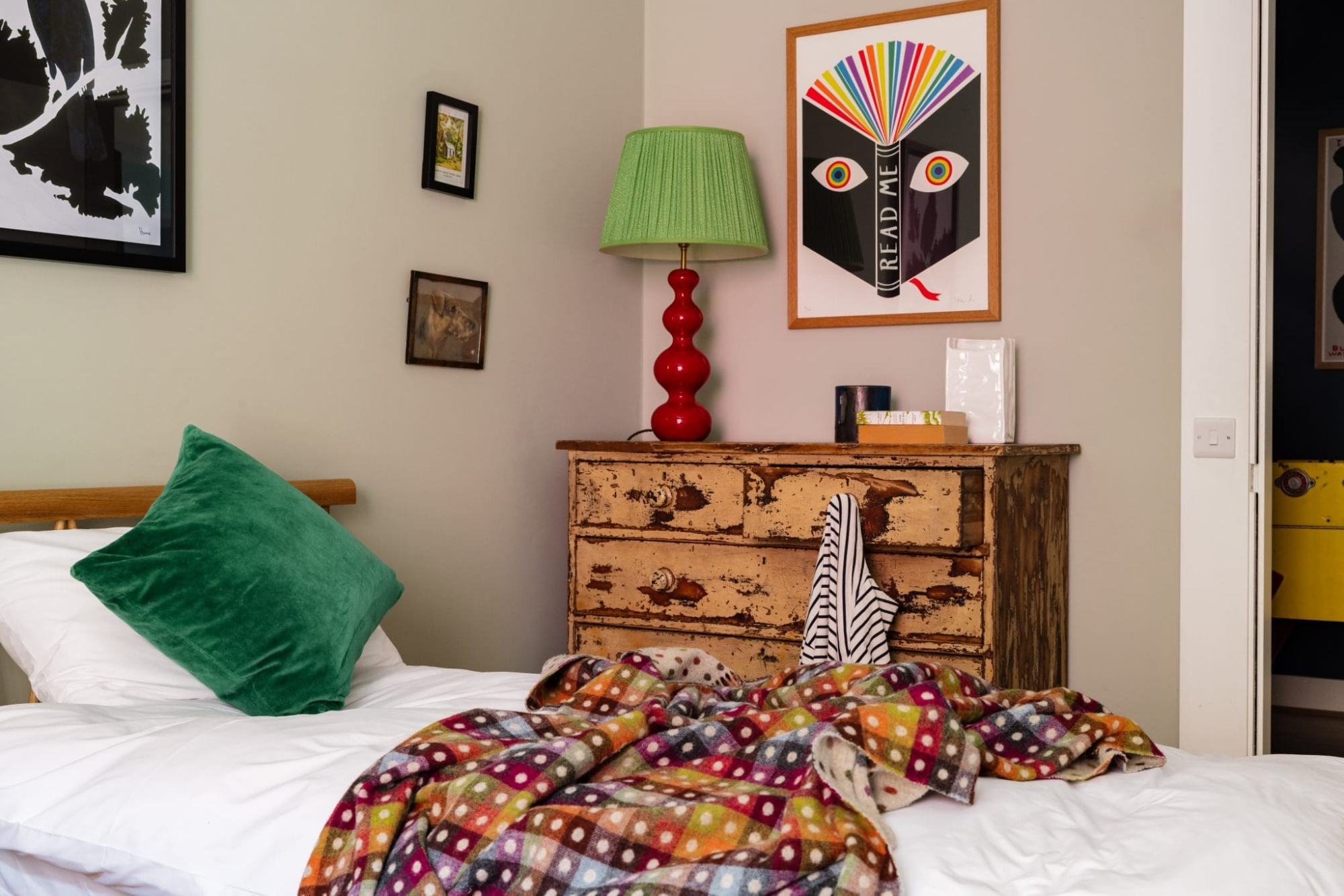 A distressed chest of drawers topped with a red table lamp with a green shade stands beside a bed with a green velvet cushion and a colourful patterned quilt.
