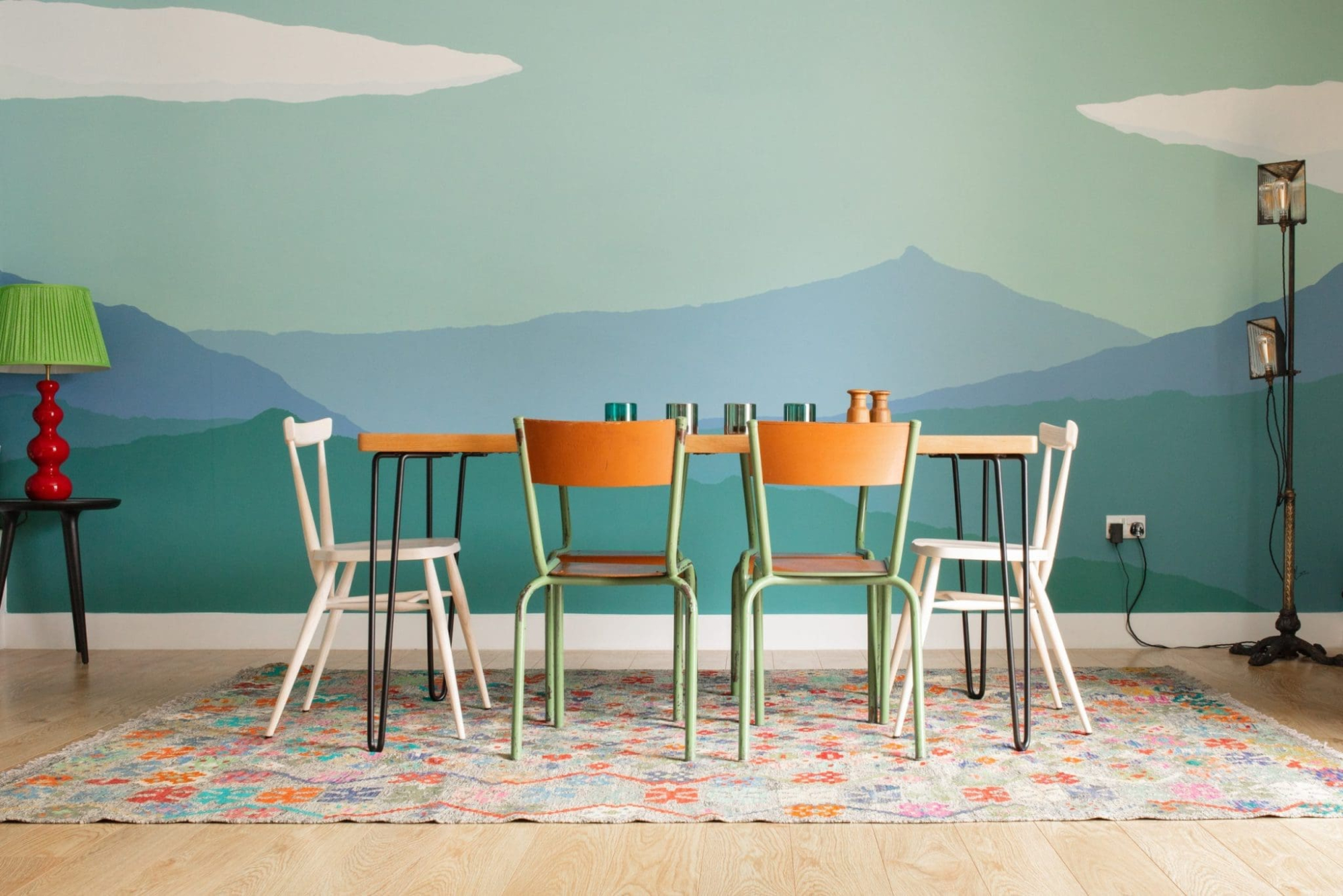 A dining area painted with a green and blue mountain mural has a wooden table surrounded by mismatched white, orange and mint metal-framed chairs, a patterned rug underfoot and a red-shaded lamp on a side table.