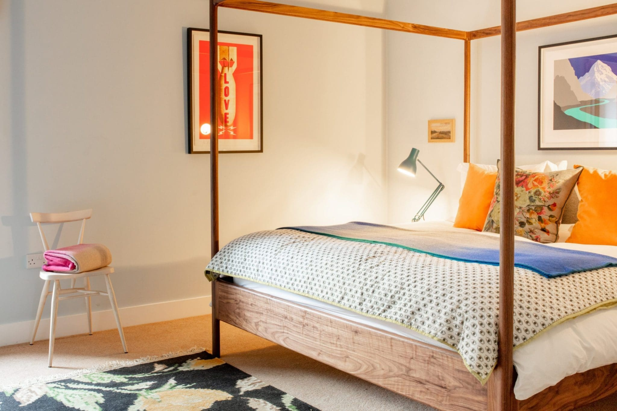 A dark wood four-poster bed is dressed in a blue, white and orange patterned duvet with scatter cushions, a teal reading lamp fixed to the wall, framed prints above the headboard and a cream chair with a pink cloth draped over it nearby.