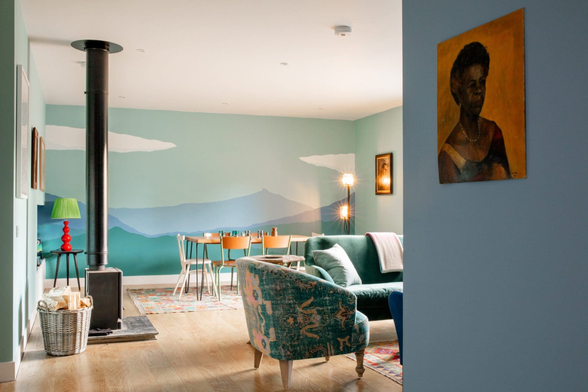 A living room has a wood-burning stove with a black flue pipe, a green sofa and a floral wingback armchair against a mint mural wall, with a red side table, a log basket and a portrait painting visible through a doorway beyond.