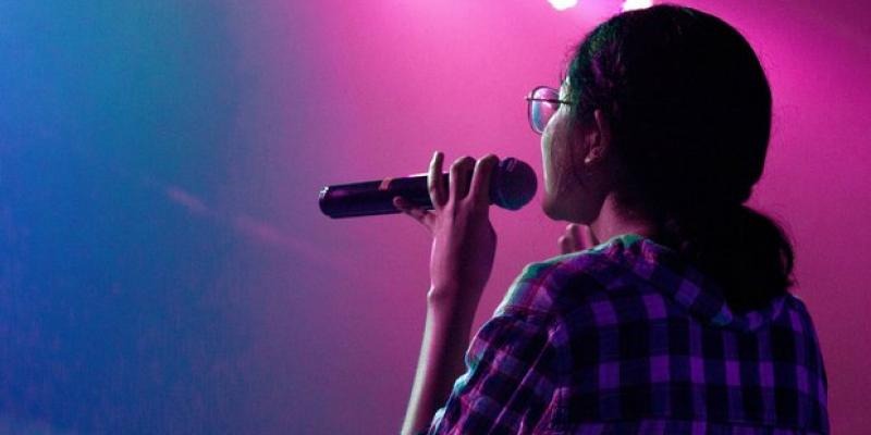 Woman in a checked shirt holding a microphone under pink stage lighting