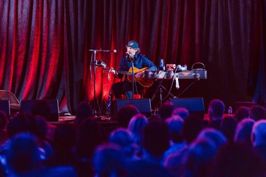 Guitarist in a cap performs before a seated audience against red curtains