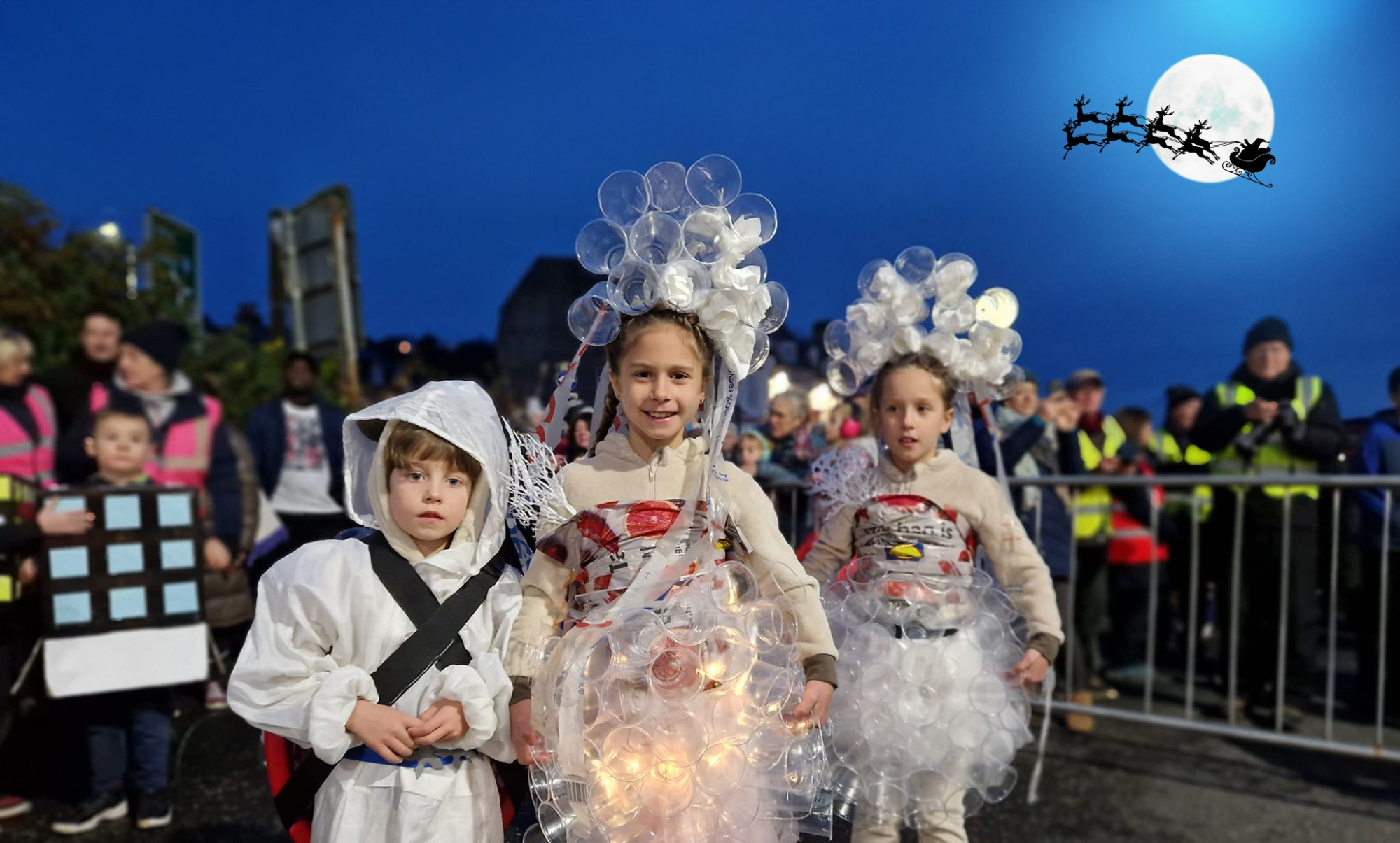 Children in illuminated costumes walk a torchlit parade beneath a Santa sleigh silhouette