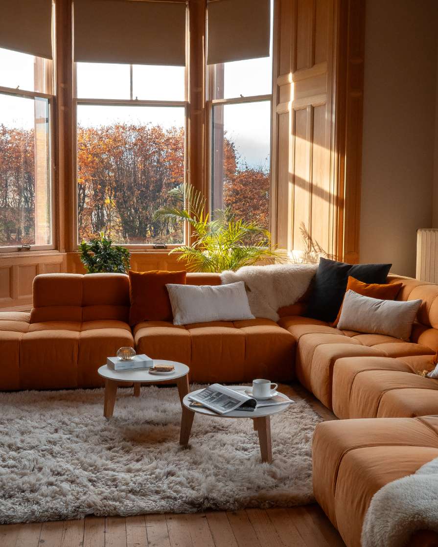A low orange sectional sofa with patterned cushions curves around a sheepskin throw and nested coffee tables, a bay window with potted plants behind.