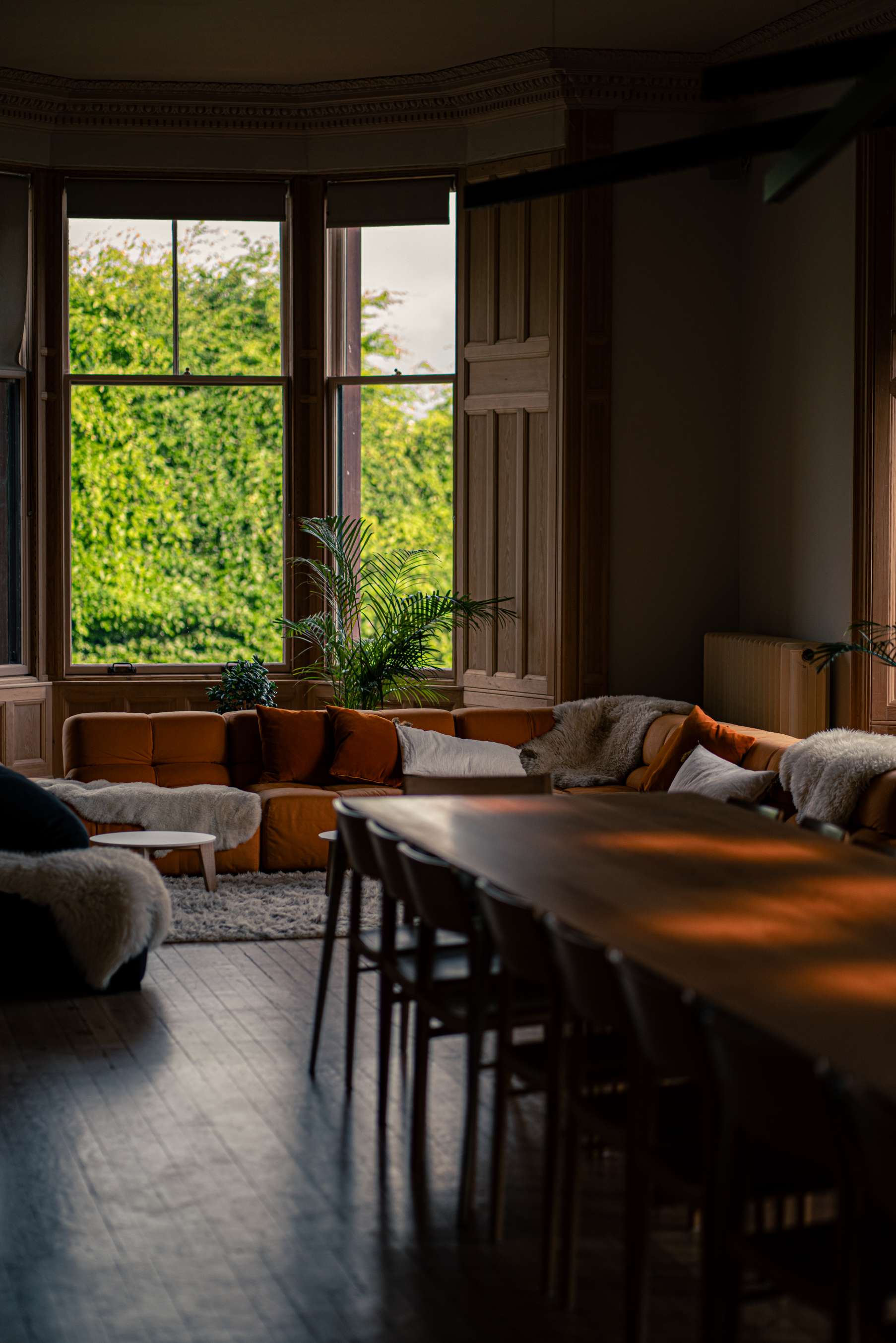A long wooden dining table and chairs are seen from a doorway, an orange sofa and windows visible in the room beyond.