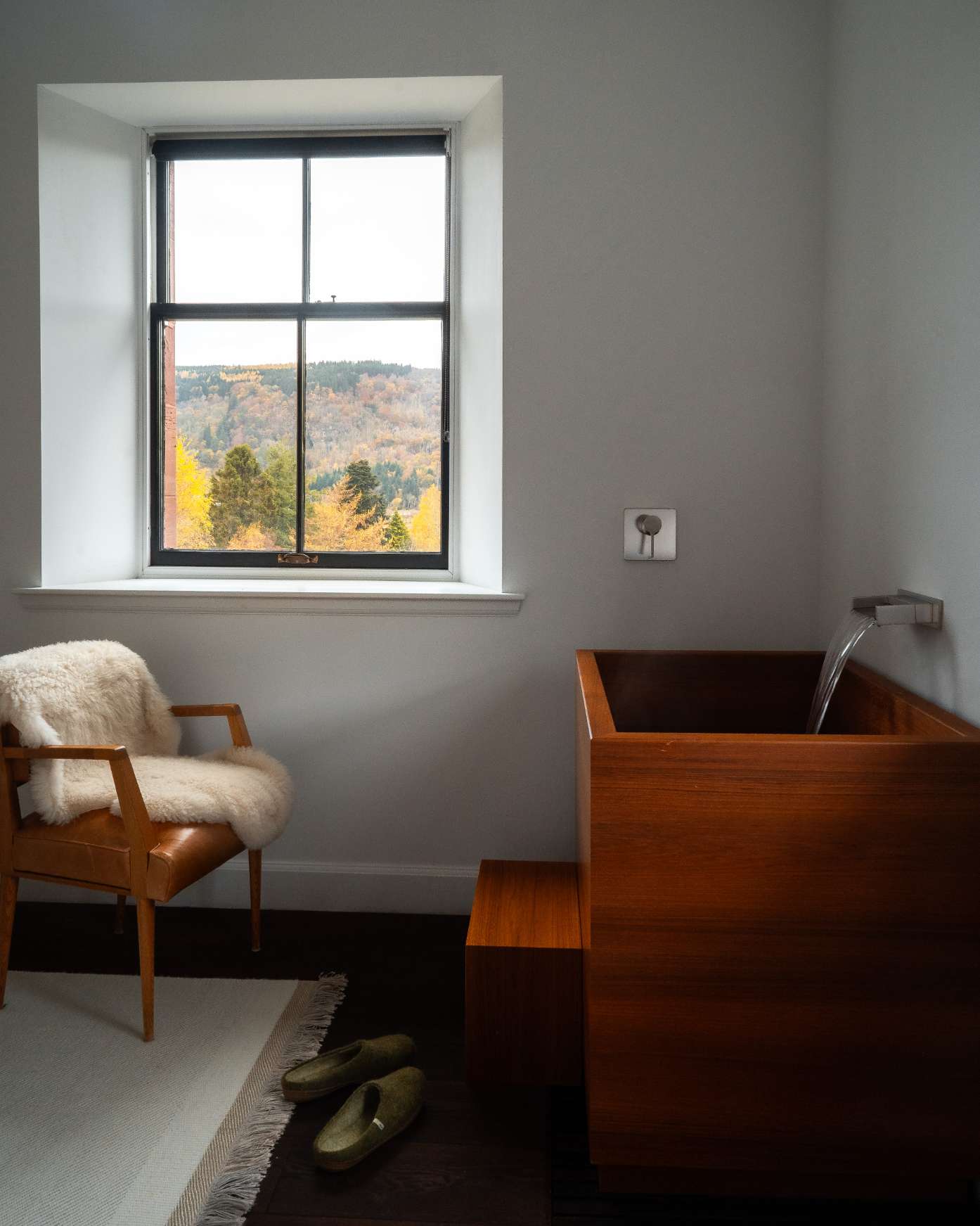 A dark wood bath stands beneath a window looking out to autumn-coloured hills, a sheepskin-draped leather armchair and a pair of slippers nearby.