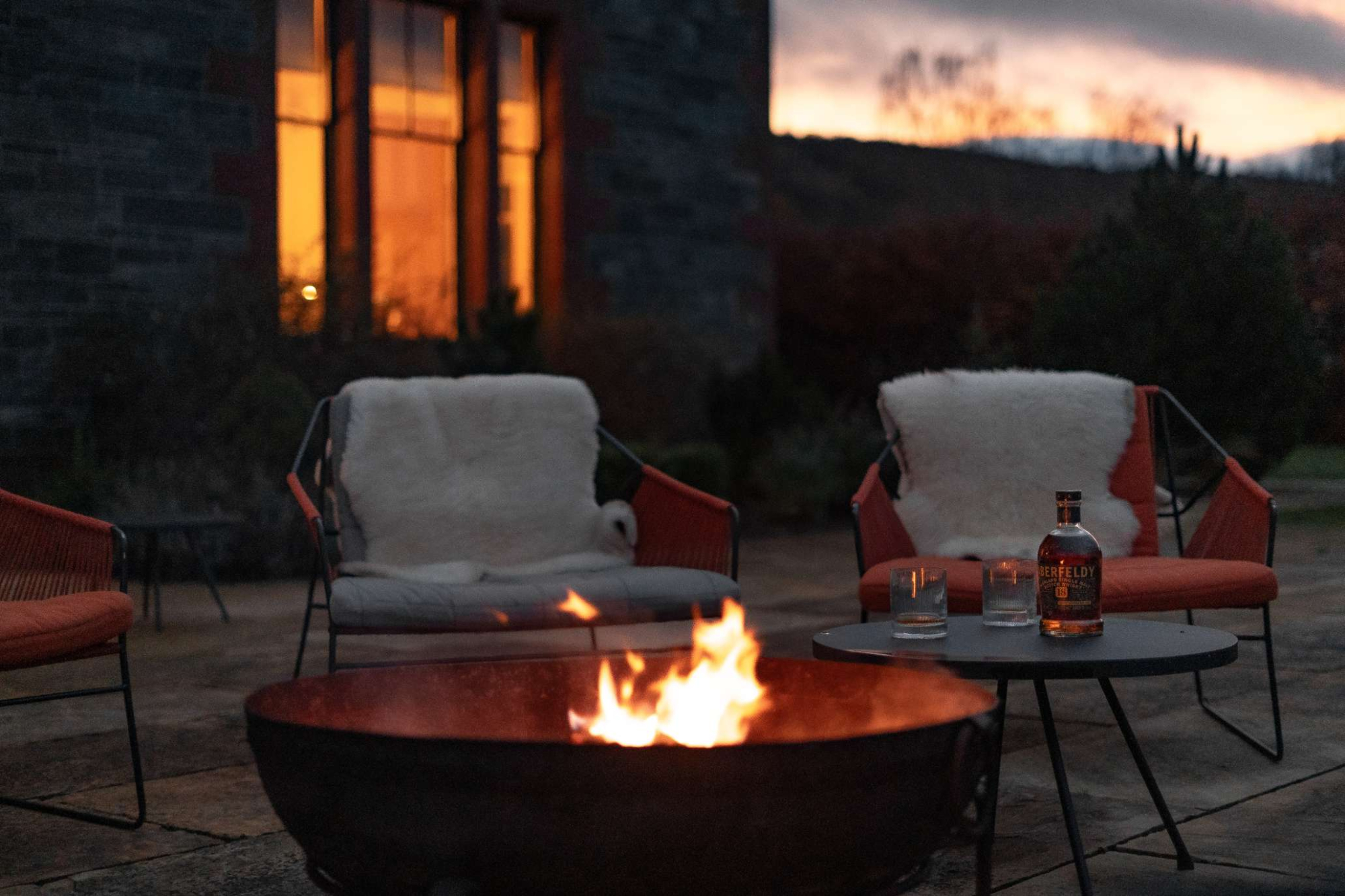 Flames rise from a firepit between sheepskin-draped chairs at dusk, a bottle and glasses set on a small table beside the lit windows of the house.