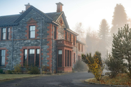 A red sandstone and grey stone Victorian house stands wreathed in morning mist, a shrub with orange autumn leaves in the foreground.